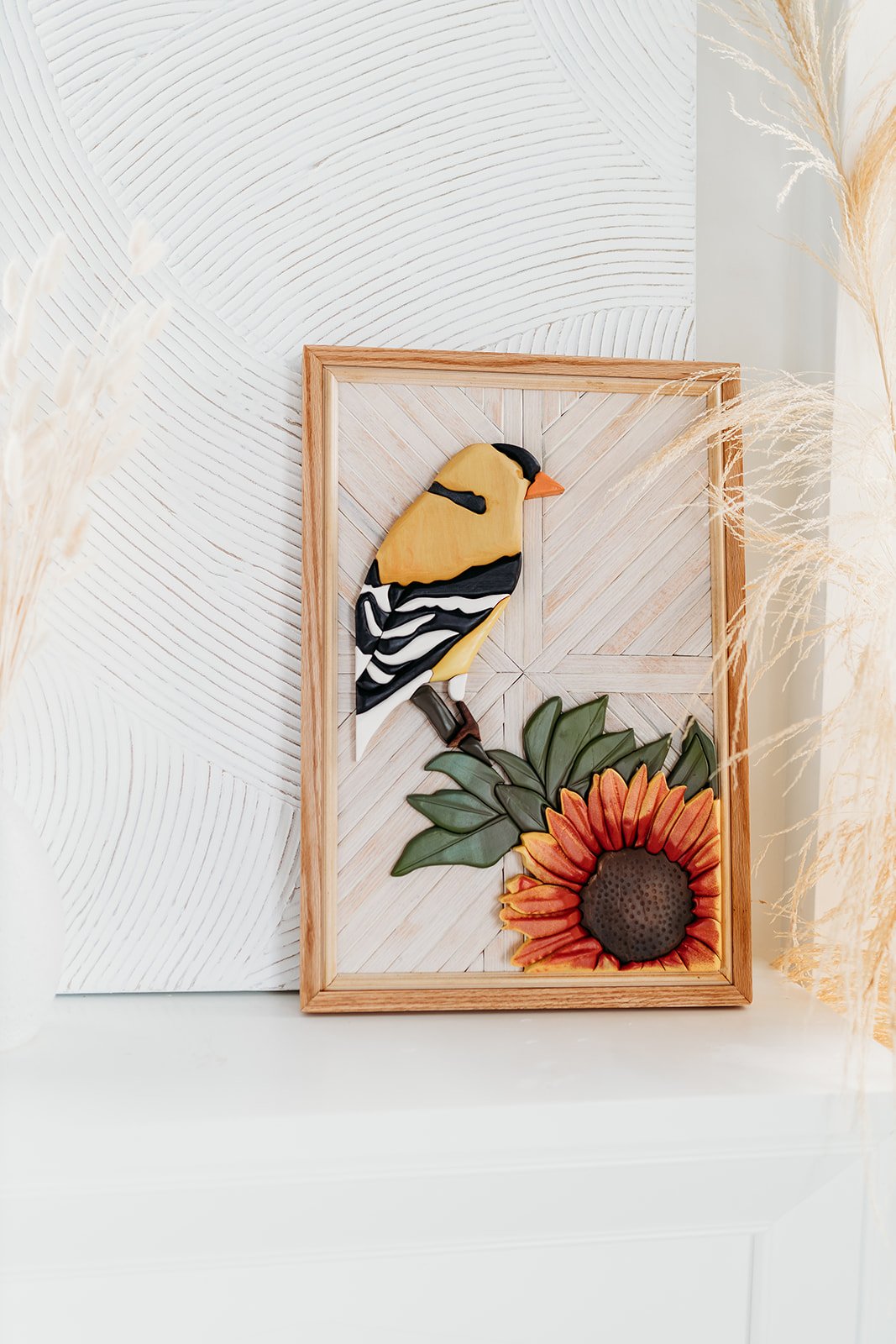 Wooden framed artwork featuring a yellow bird and a sunflower on a patterned white background, with decorative grasses surrounding the frame.