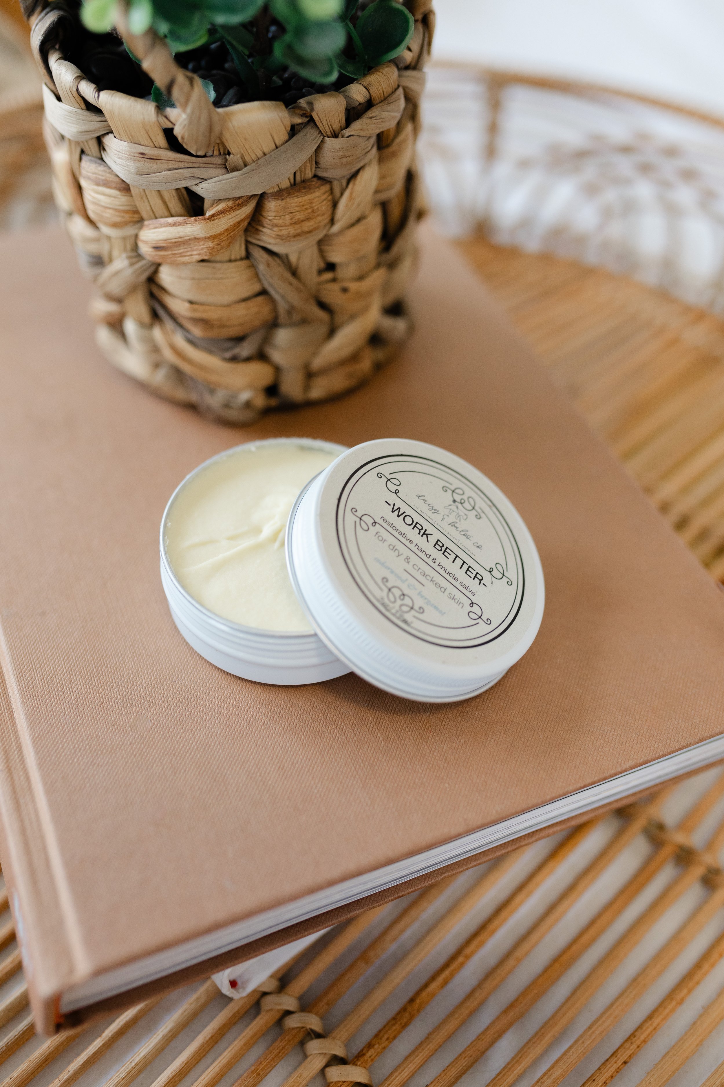 Tin of hand cream labeled 'Work Better' on a beige notebook next to a woven basket with a plant.