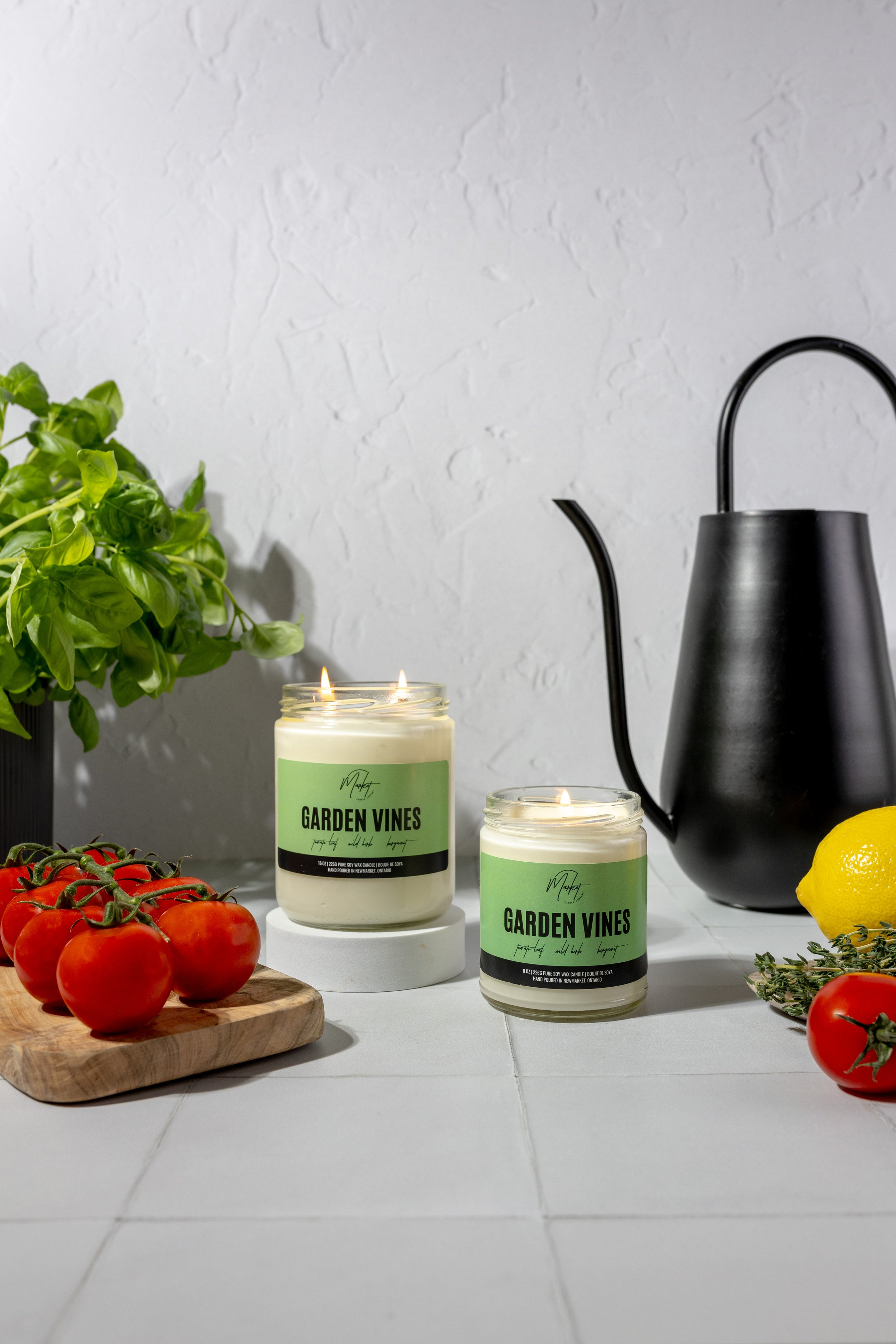 Two "Garden Vines" candles on a table with tomatoes, basil, lemon, thyme, and a black watering can on a gray background.