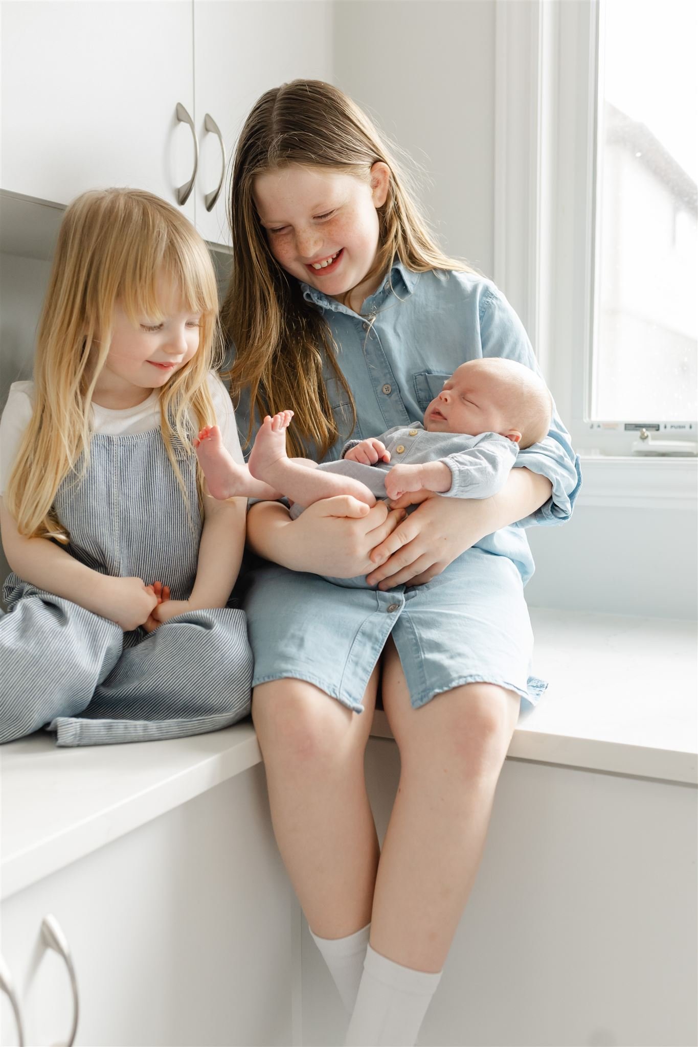 Two young girls sitting on a white countertop, one holding a sleeping baby. All are wearing light-colored clothes, with the older girl smiling and the younger one looking down at the baby. Bright and natural lighting from a nearby window.