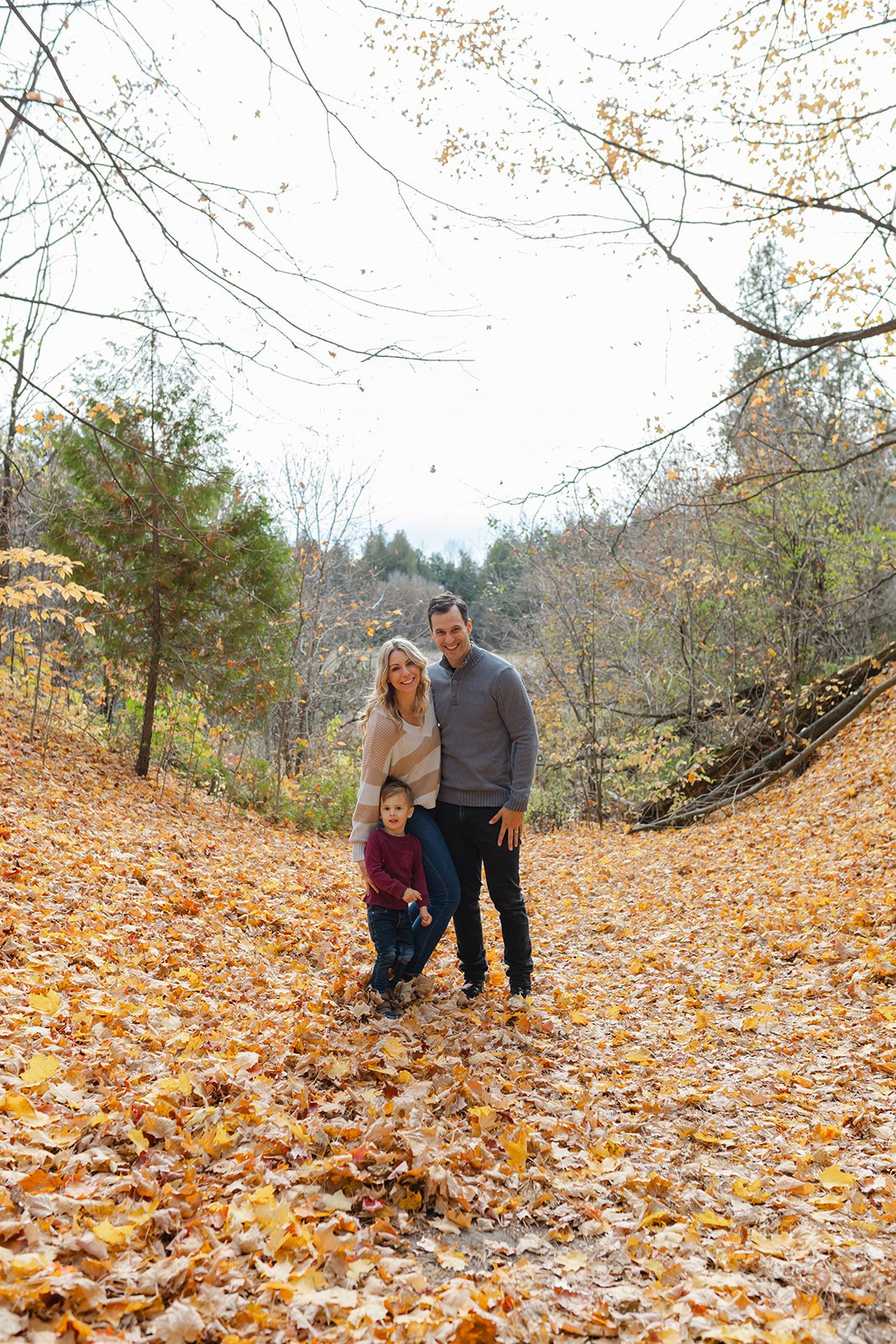 A family of three posing in a wooded area covered with autumn leaves. The parents are standing with a child in front, surrounded by trees with fall foliage.