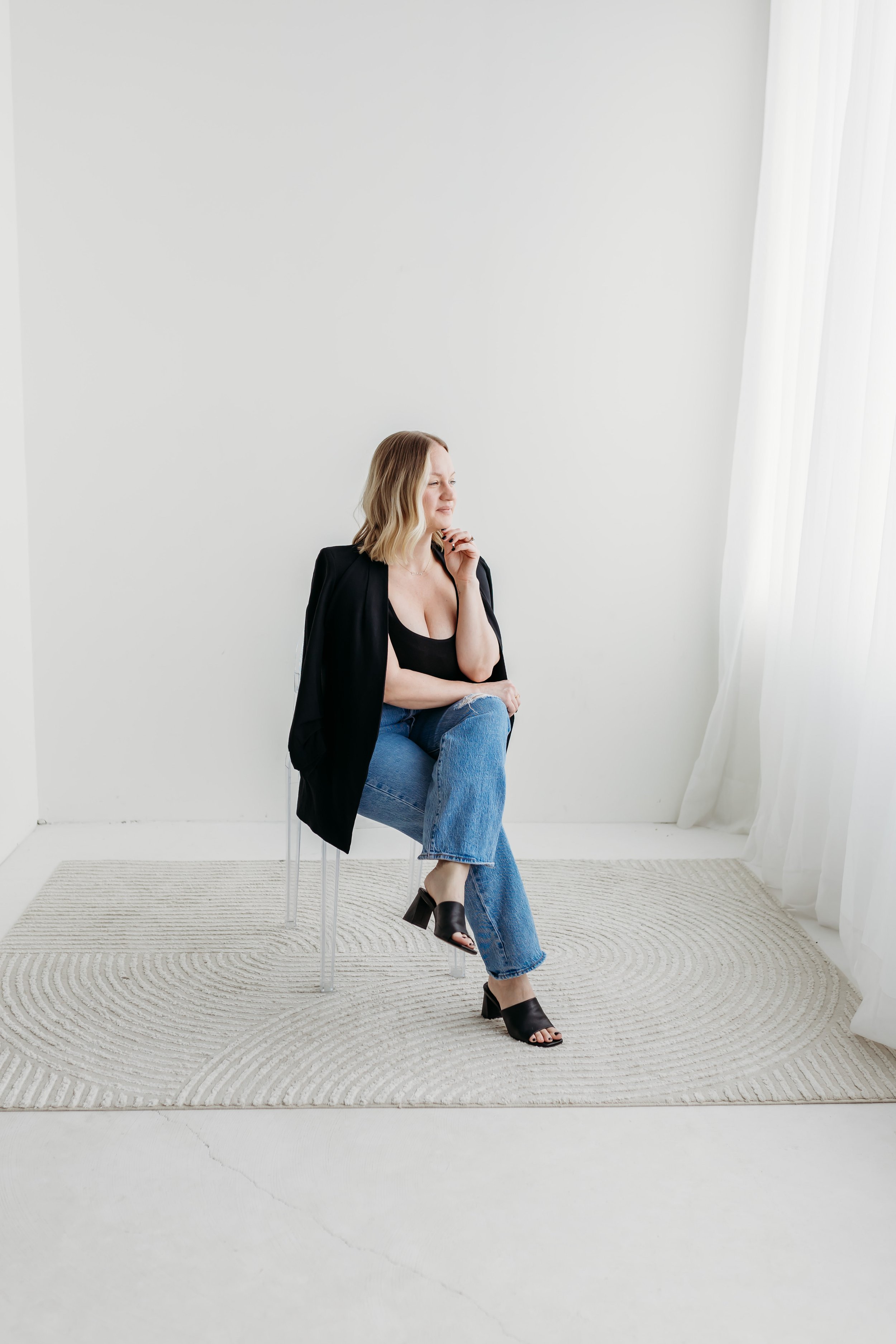 Woman sitting on a chair, wearing a black shirt, blazer, blue jeans, and black heels in a minimalist room with white walls and curtains.