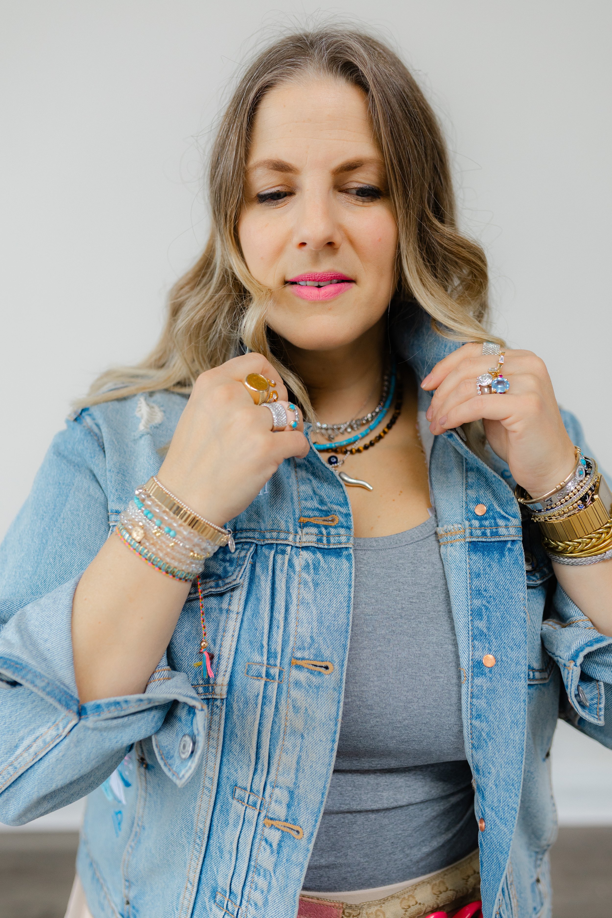 Woman wearing a denim jacket, gray tank top, and multiple jewelry pieces including bracelets, rings, and necklaces, with long light brown hair.