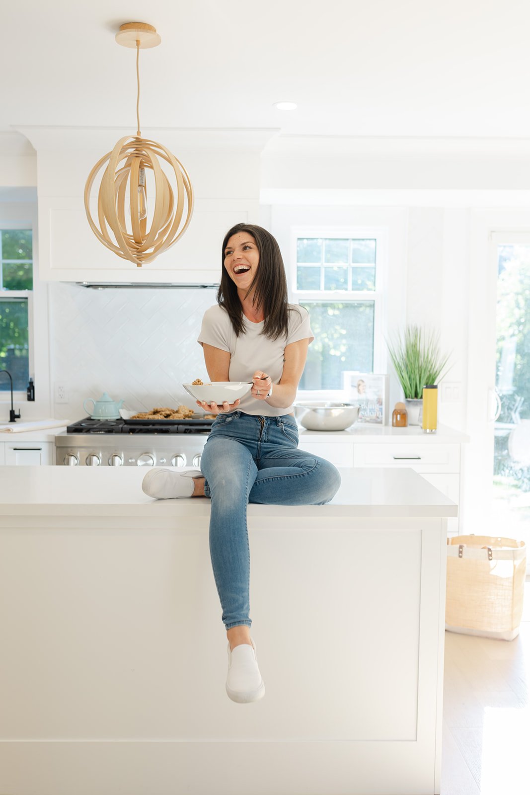 A woman sitting on a kitchen counter, holding a bowl, smiling, wearing a white T-shirt and jeans. The kitchen is bright with a modern design, featuring a hanging light fixture and a stove in the background.