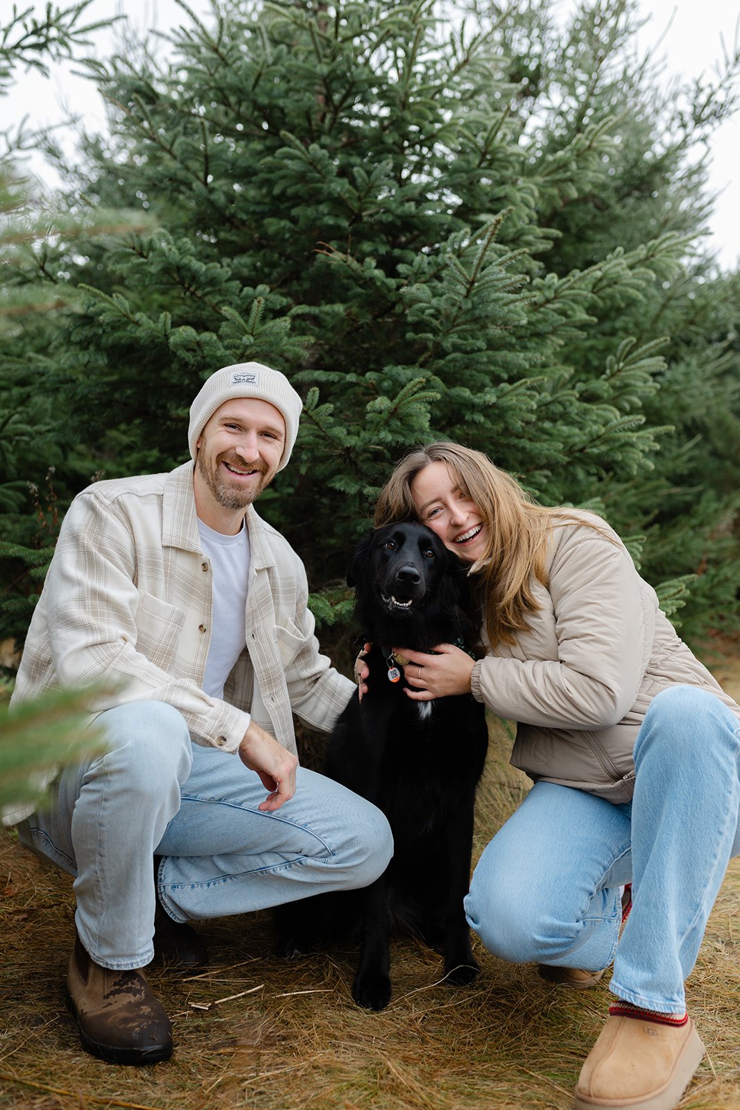 A smiling couple kneeling next to a black dog in an outdoor setting with pine trees.
