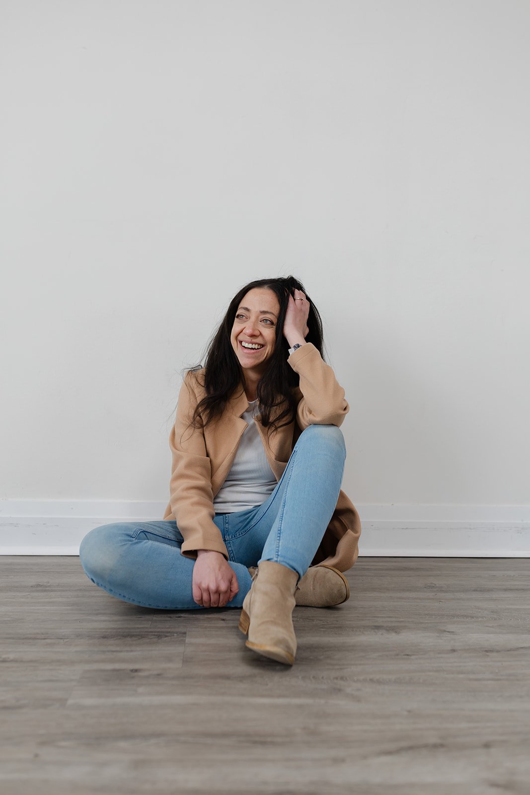 Woman sitting on floor wearing a camel coat and jeans, smiling.