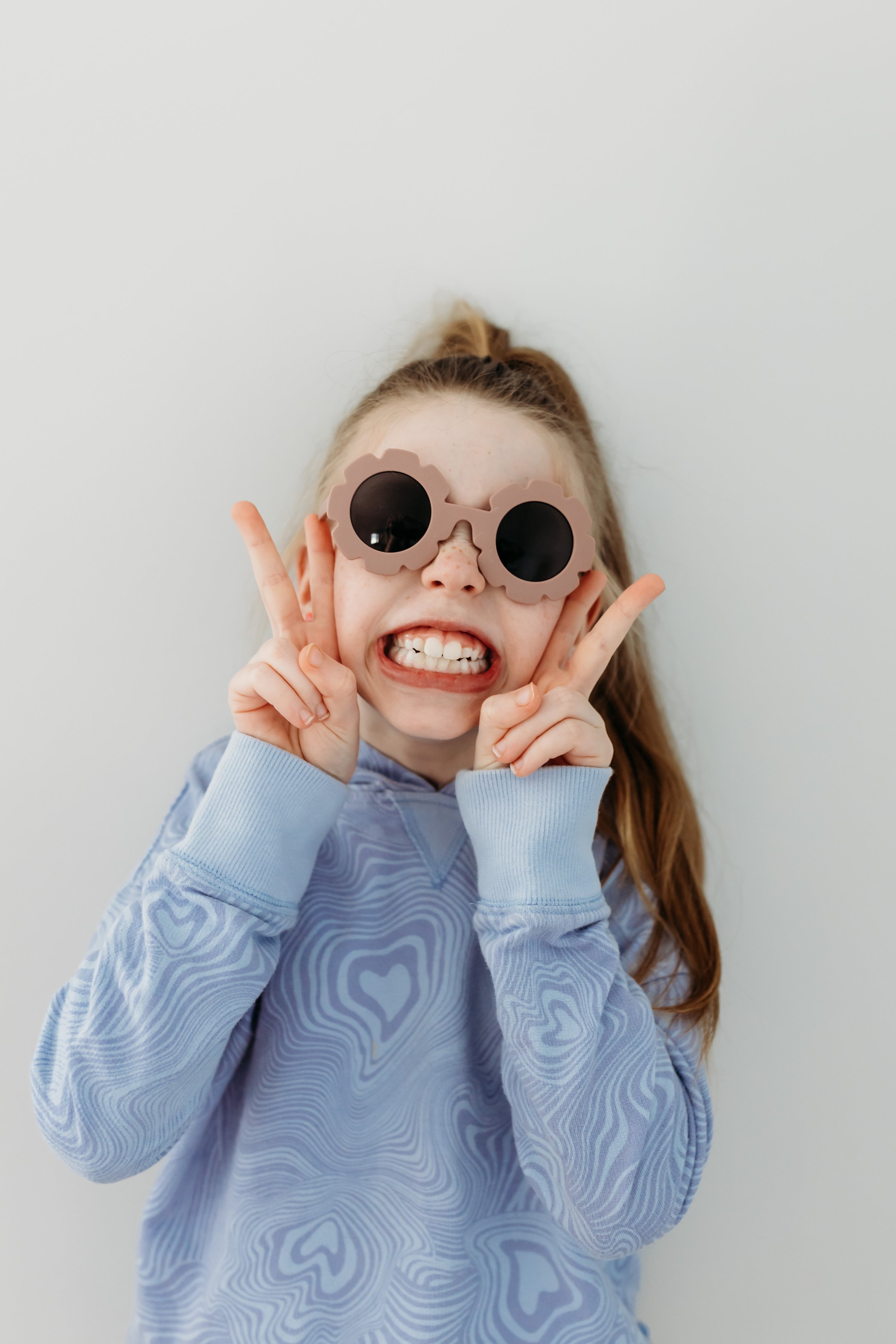 Smiling child wearing circular sunglasses and a blue patterned hoodie, making peace signs with both hands.
