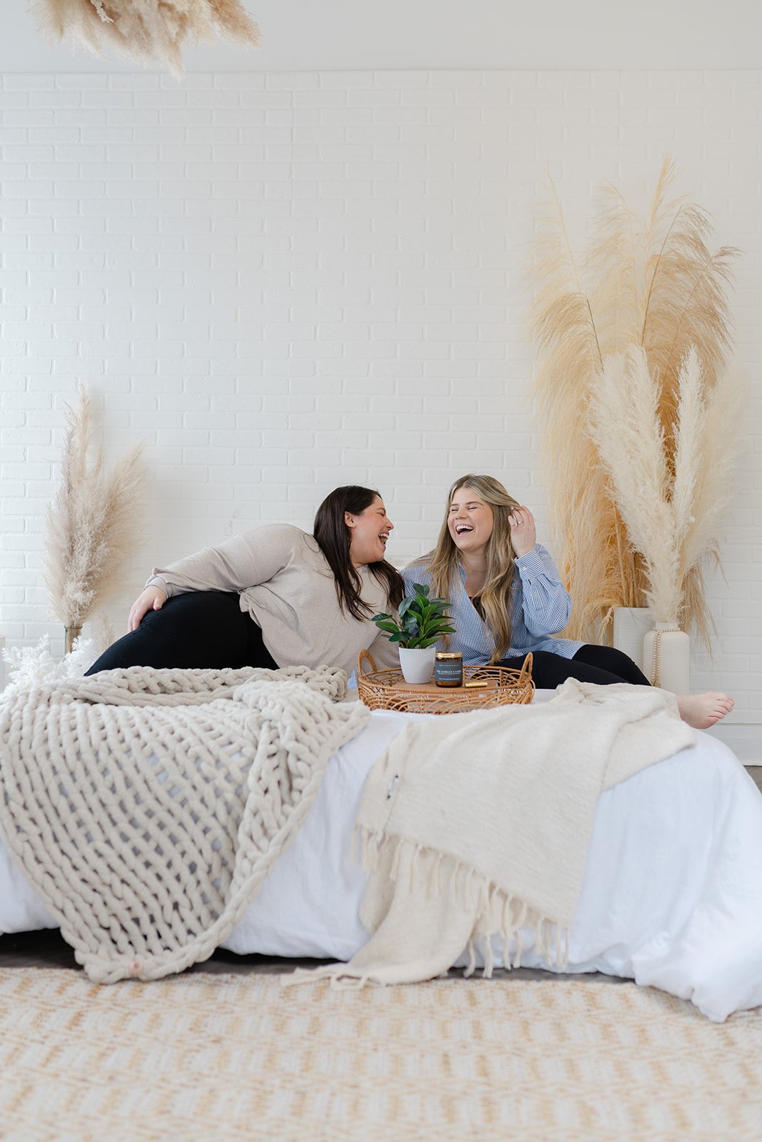 Two women sitting on a bed with a cozy beige knit blanket and a tray holding a plant and a candle, enjoying a light moment and laughing, with dried pampas grass decor against a white brick wall.