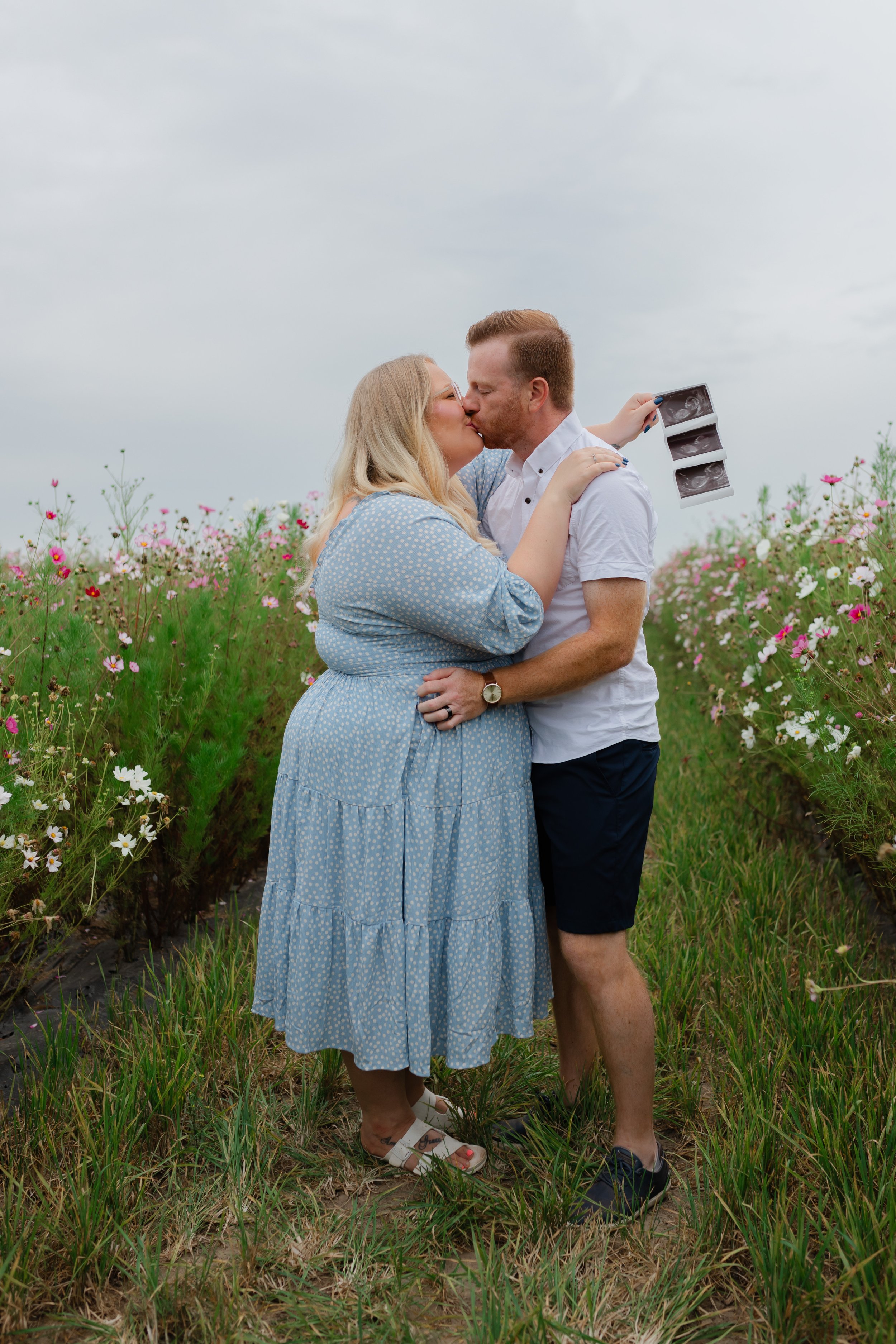 A couple is kissing in a flower field, with the woman holding an ultrasound image.