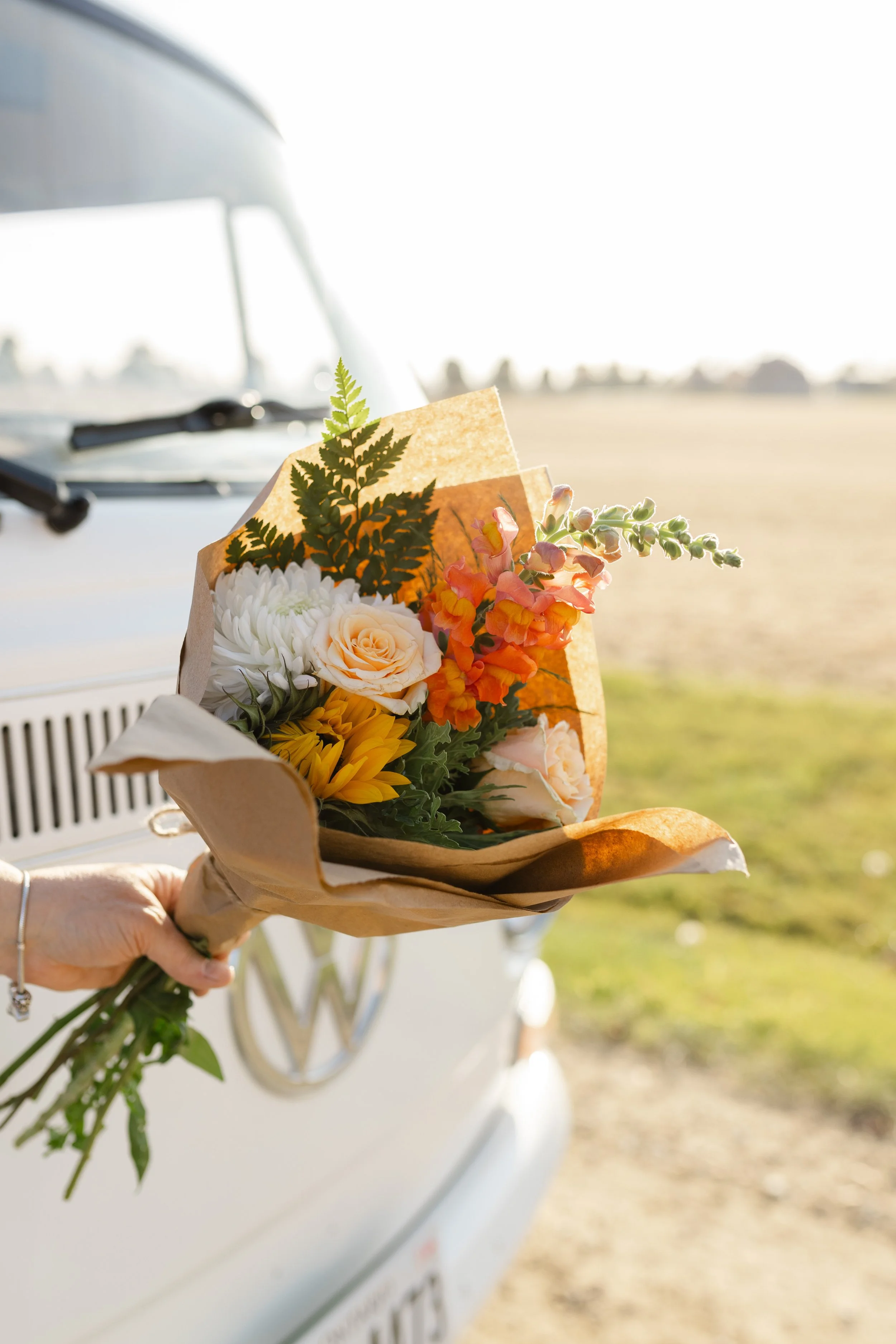 Person holding a bouquet of flowers, including roses, sunflowers, and greenery, wrapped in brown paper in front of a white vintage van, outdoors on a sunny day.