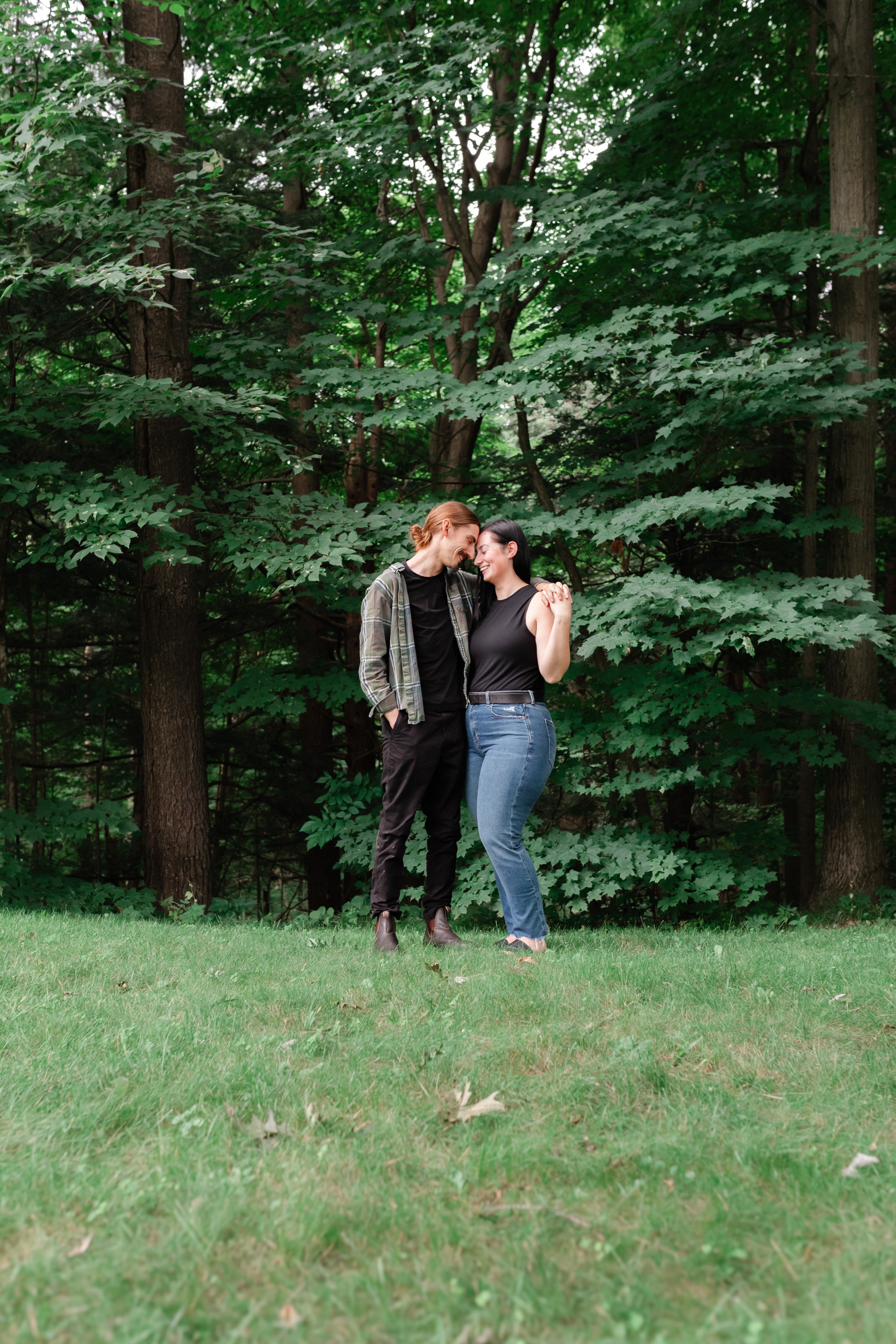 A couple standing closely together in a forested area, with trees and greenery in the background. The man is wearing a plaid shirt over a black t-shirt and the woman is wearing a black sleeveless top and jeans. Both are smiling and appear content.