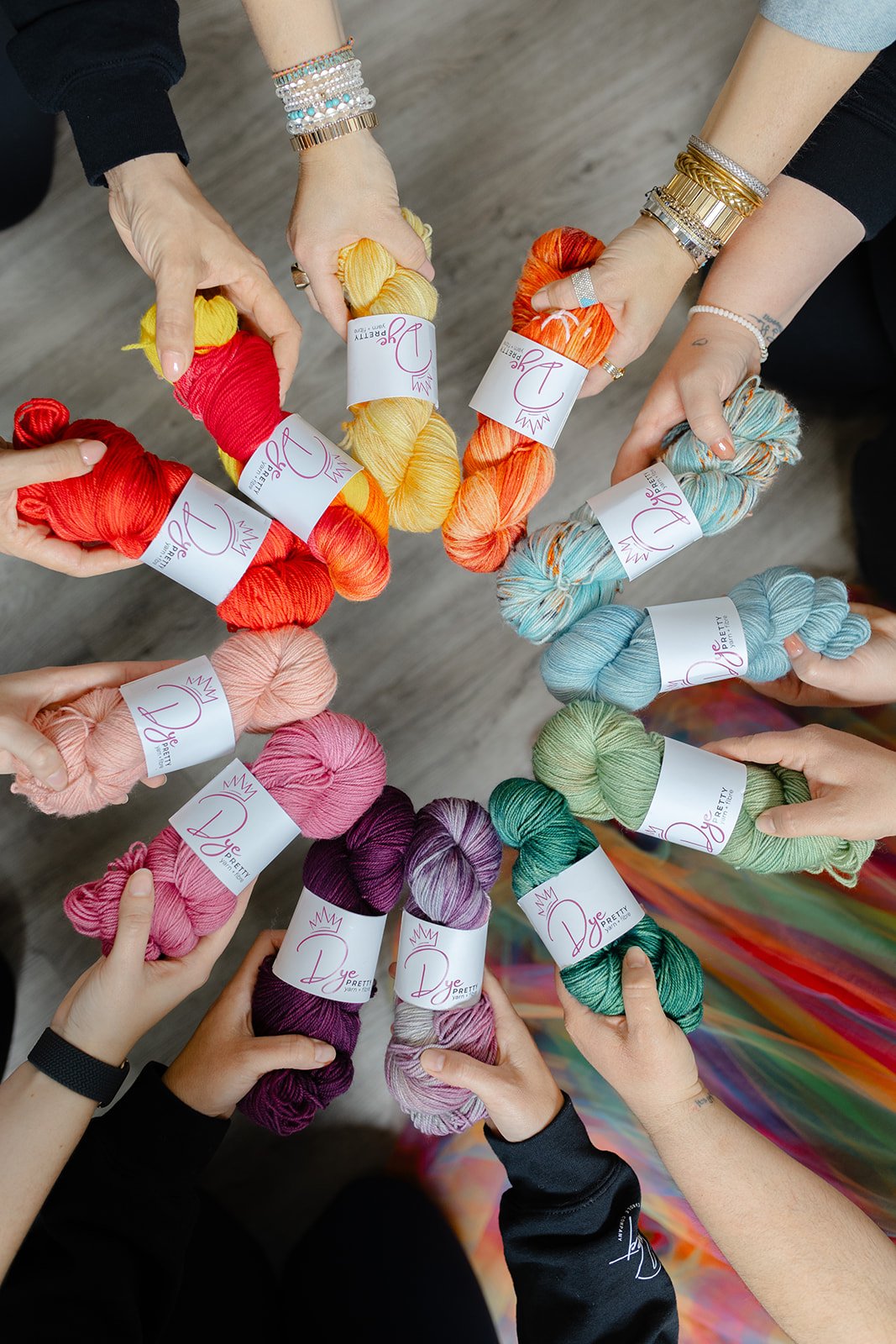 People holding colorful skeins of yarn arranged in a circle on a wooden floor.