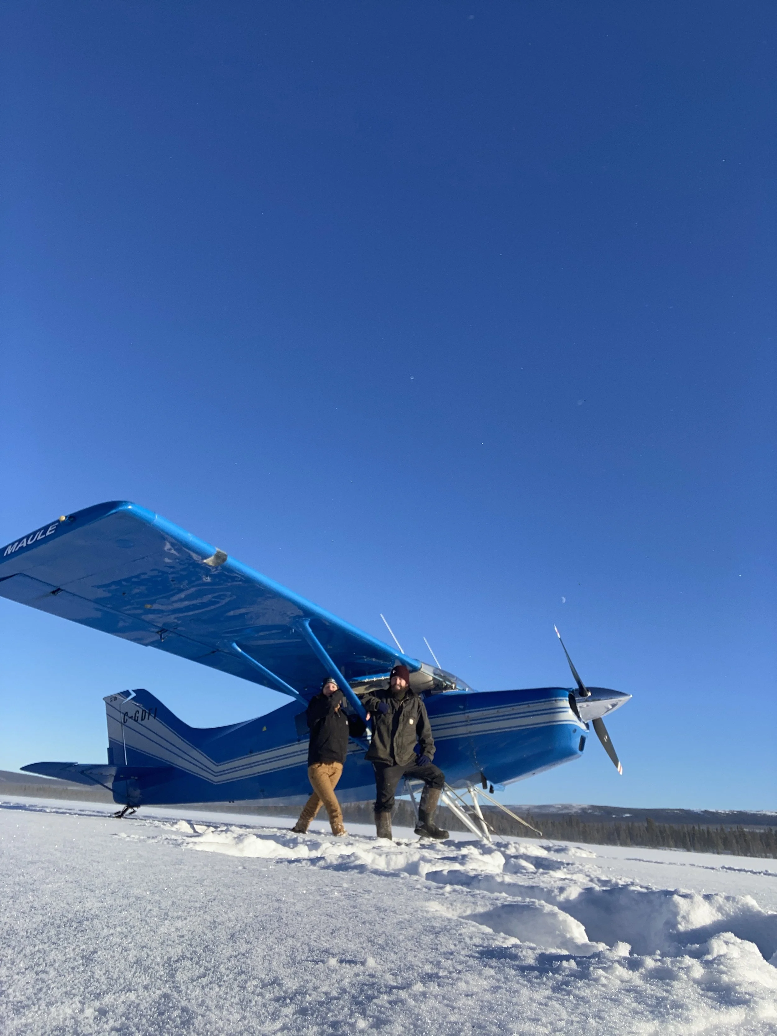 Two people standing in front of a blue propeller plane on a snowy field with a clear blue sky.