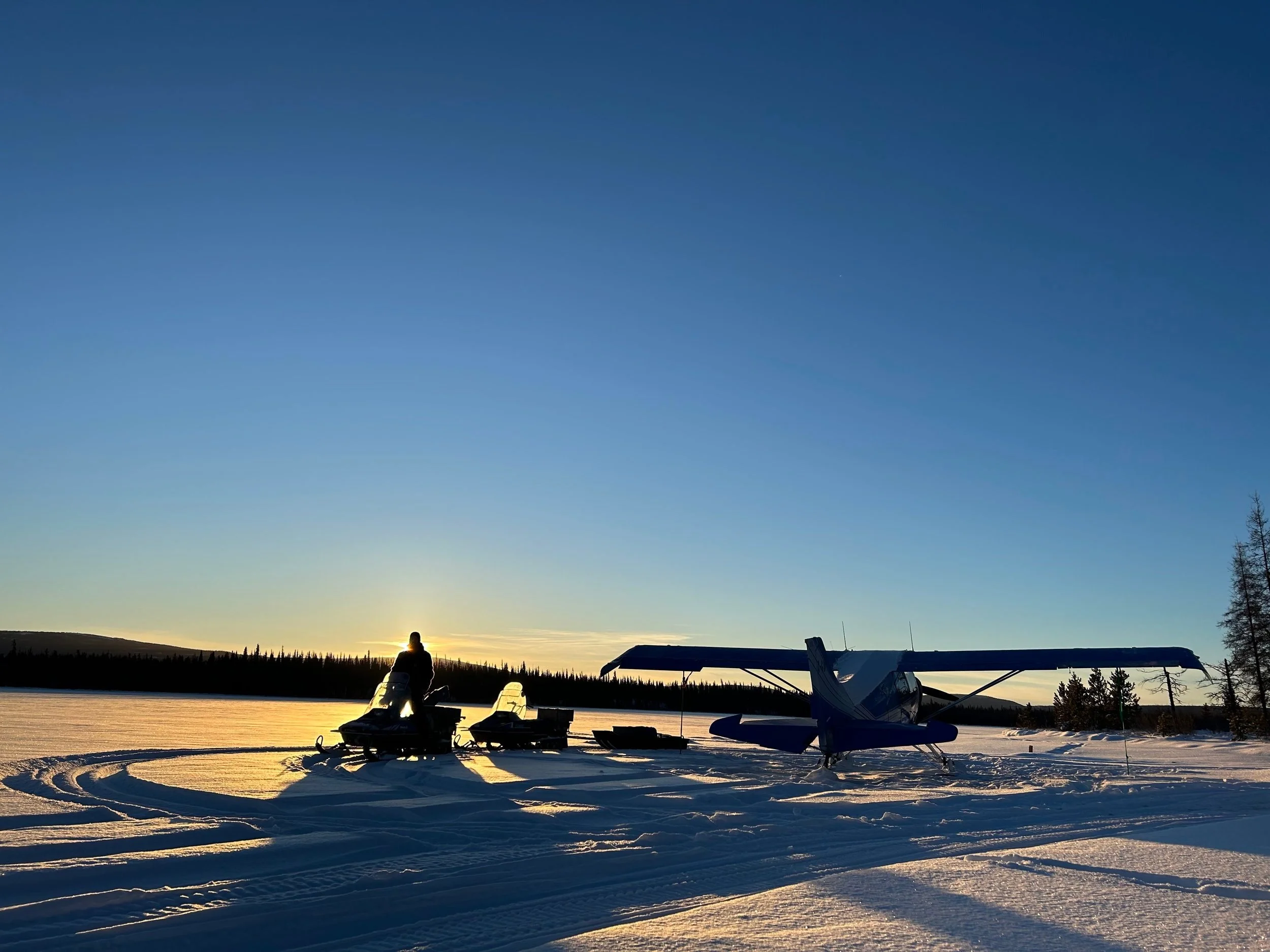 Silhouette of a person on a snowmobile and an airplane on a snowy landscape at sunset with a clear sky.