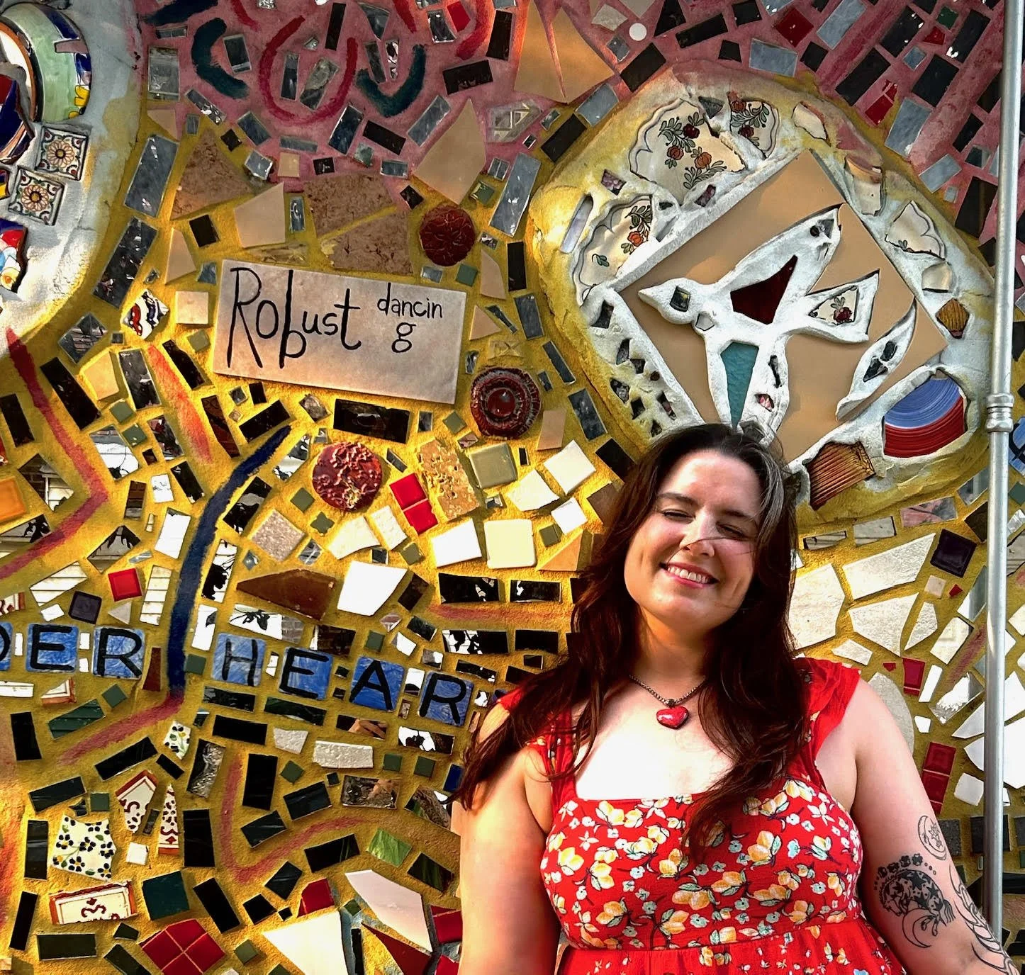 Woman with long dark hair wearing a red dress with a floral pattern and a heart-shaped necklace, smiling in front of a colorful mosaic wall.