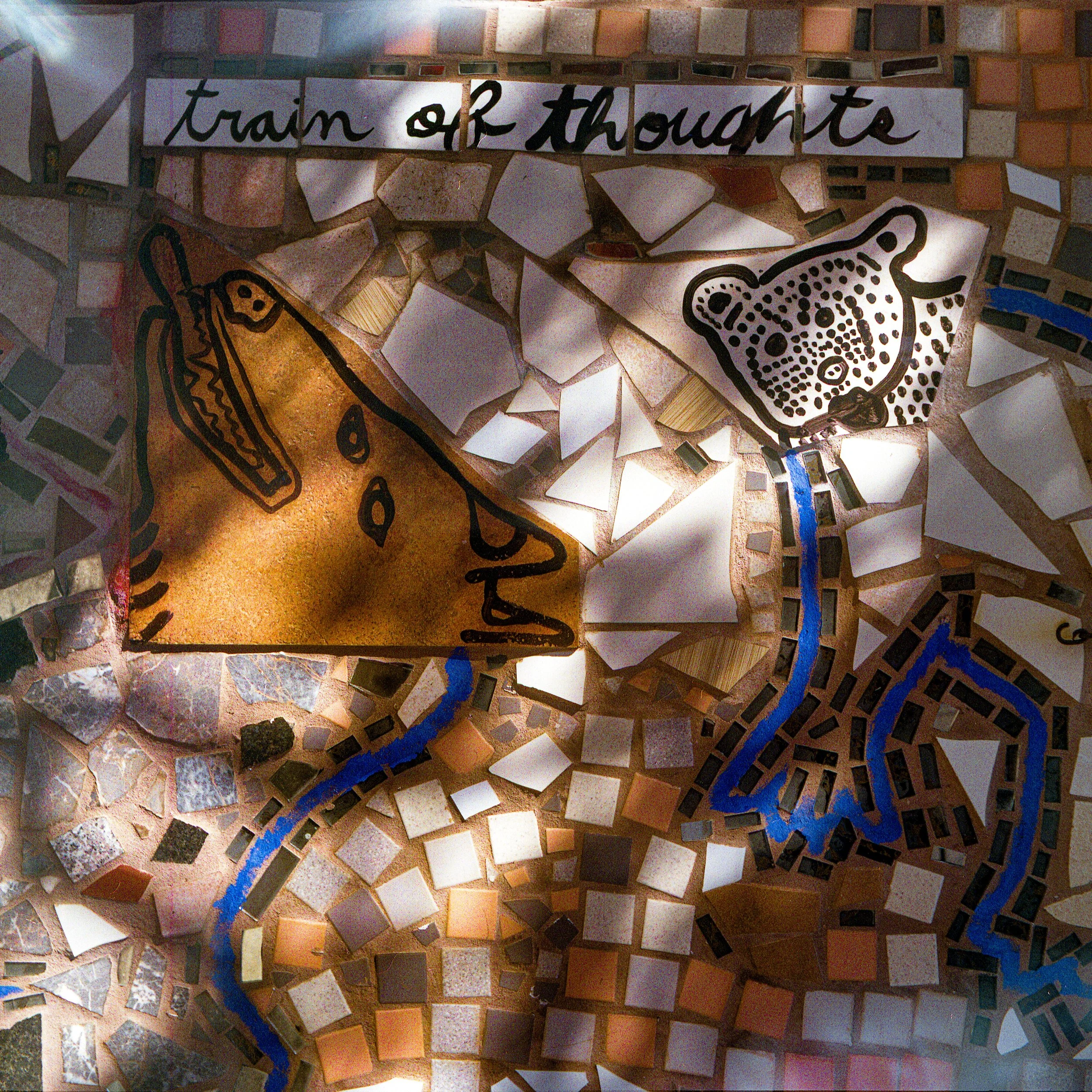 A mosaic mural with the words 'train of thought' written in black cursive letters on white tile background, featuring a black outlined leopard face on the right and other abstract shapes and colors surrounding it.