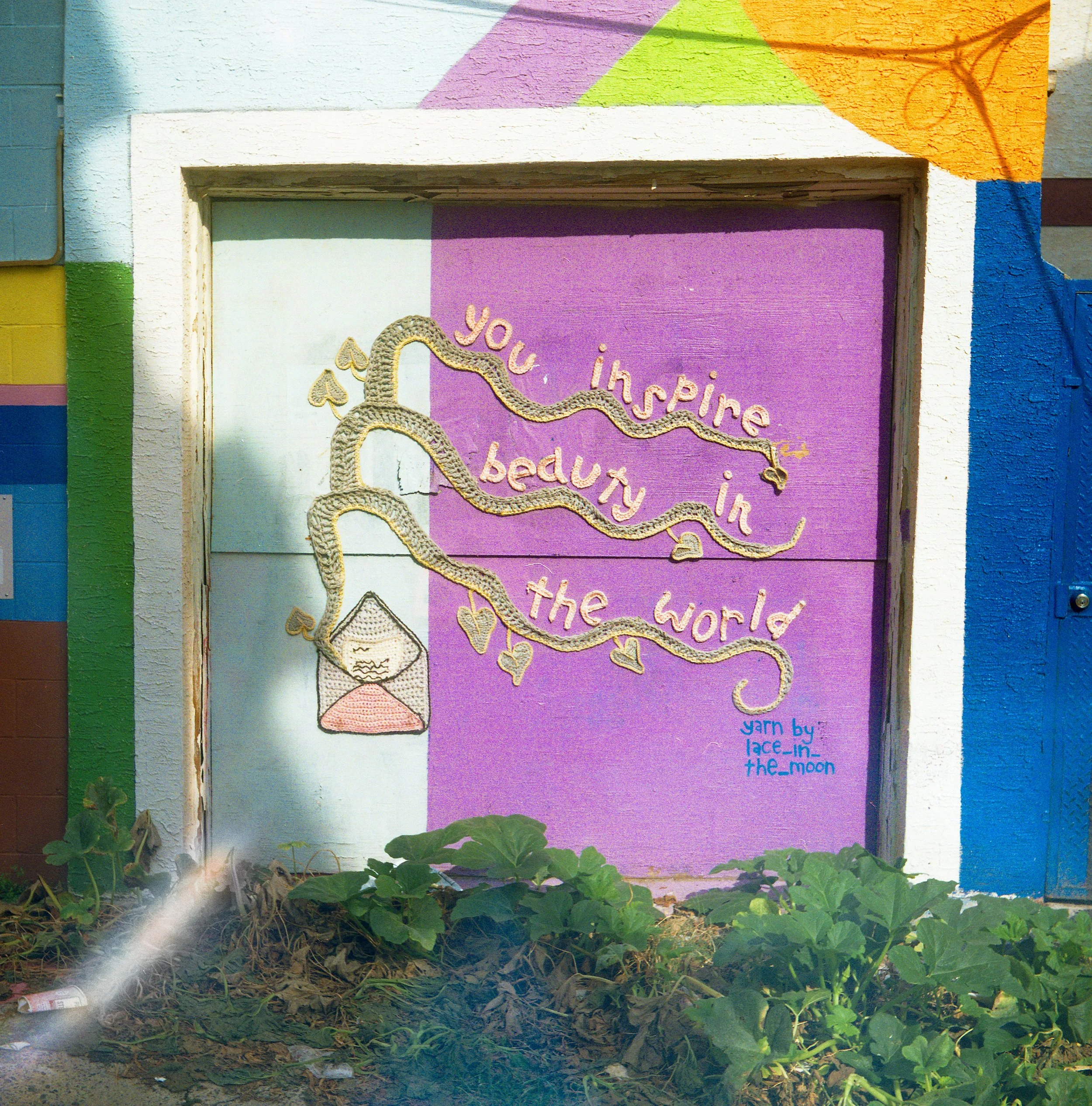 Colorful wall with painted geometric shapes and a textured sign that reads 'you inspire me in the world' with a knitted snake decoration. Green plants and dirt are at the base of the wall.