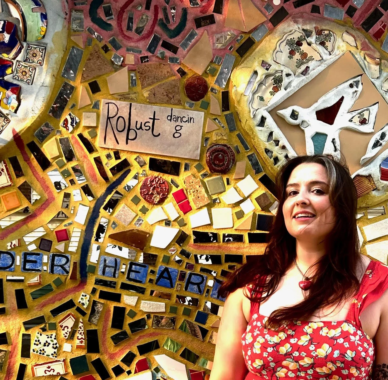 A woman with long dark hair wearing a red floral dress, standing in front of a colorful mosaic artwork with various tiles, shapes, and patterns, including a sign that reads 'Robust dancing' and part of the phrase 'PER THE ART.'