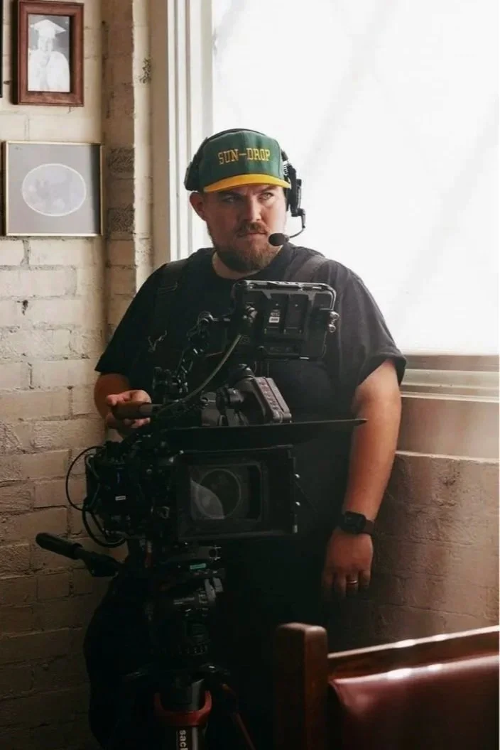 A man with a beard, wearing a green and yellow cap with 'Sun-Drop' written on it, a black T-shirt, and a headset with a microphone. He is operating a professional video camera in a room with a white brick wall and a window, with a framed photo on the wall behind him.