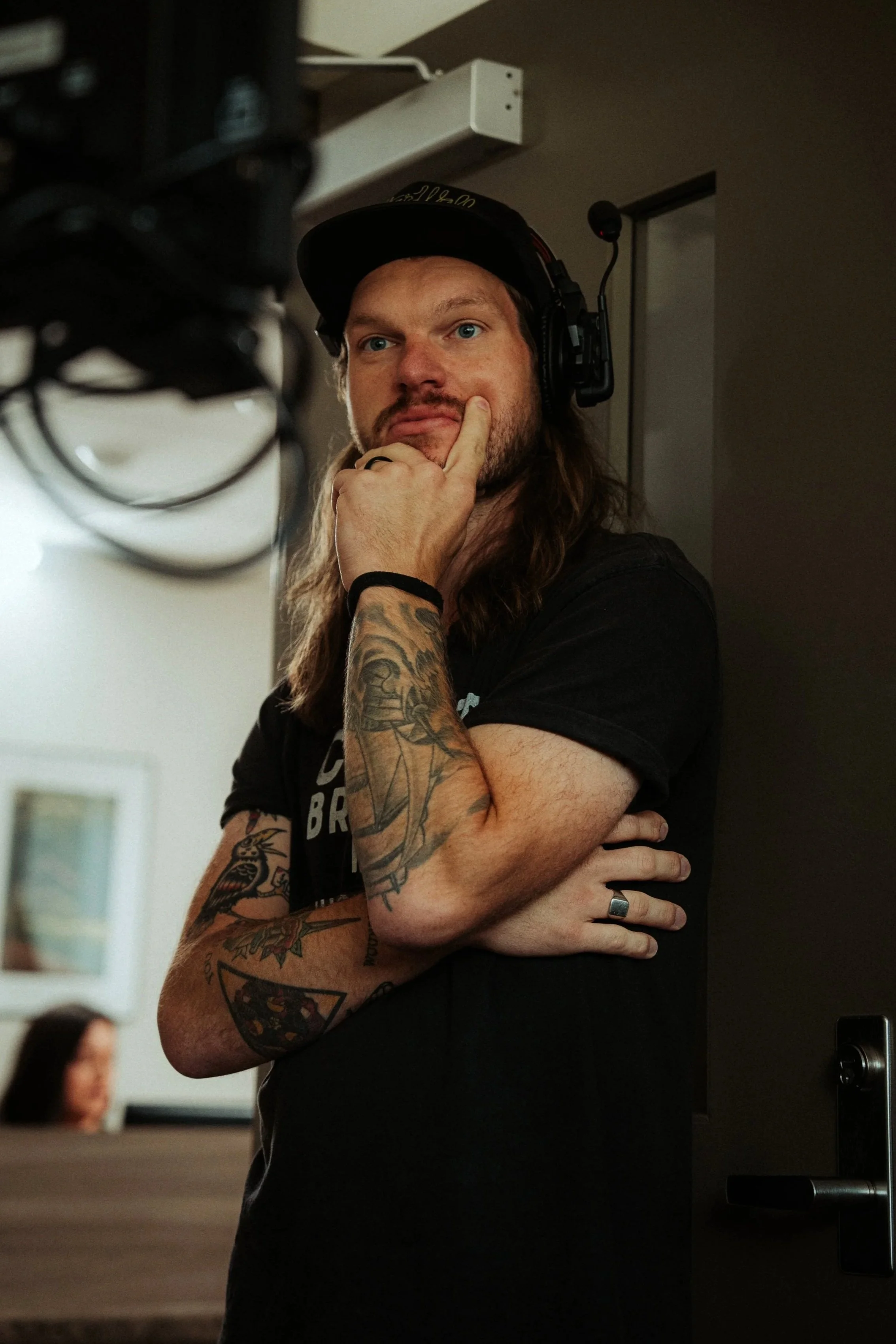A person with long hair and tattoos on their arms, wearing a black hat and headphones, standing indoors with a thoughtful expression, with others blurred in the background.