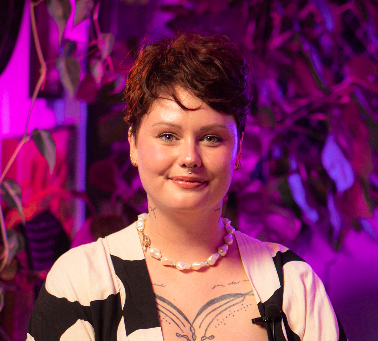 Pessoa em primeiro plano com cabelo curto, colar de pérolas e tatuagens visíveis, fundo com luzes roxas e plantas.