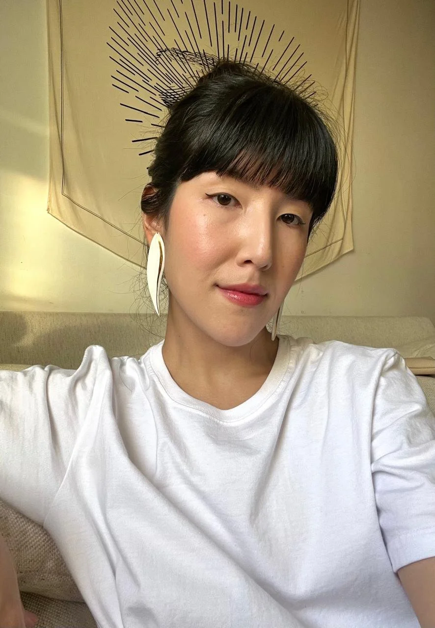 Pessoa com cabelo preto e franja, usando camiseta branca e brincos grandes, sentada em um sofá com uma peça decorativa de parede ao fundo.