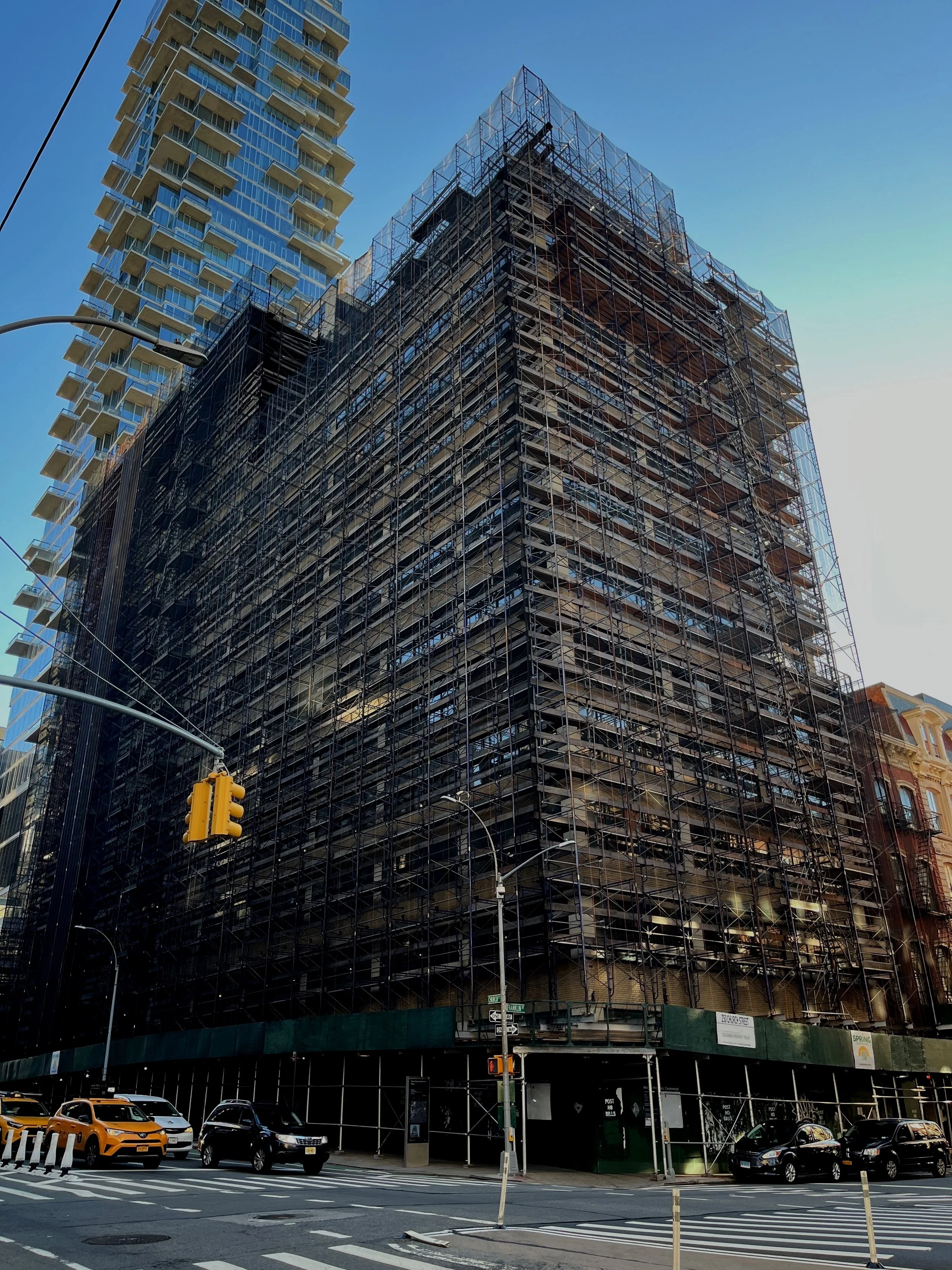 A high-rise building under construction with scaffolding on all sides, situated on a city street with cars and traffic lights.