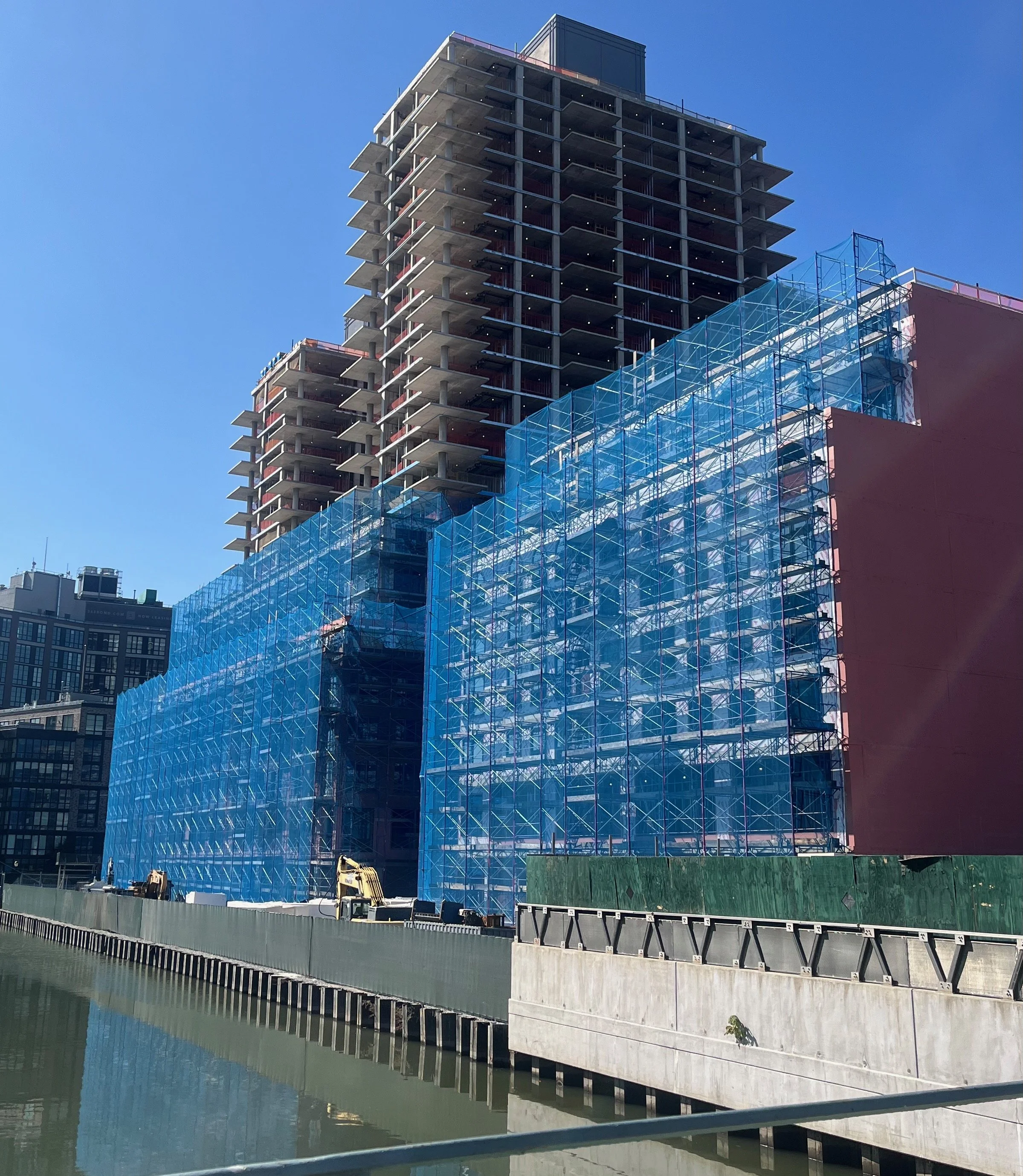Under construction high-rise building with exposed concrete floors and blue safety netting on a waterfront during daytime.