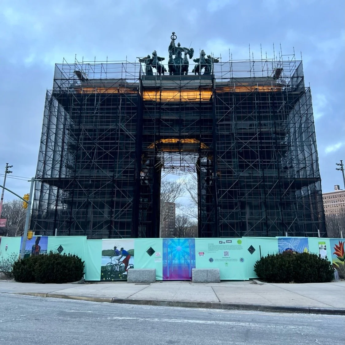 The Arc de Triomphe under renovation with scaffolding covering the monument, and it is behind a construction fence with colorful banners. Trees and buildings are visible in the background.