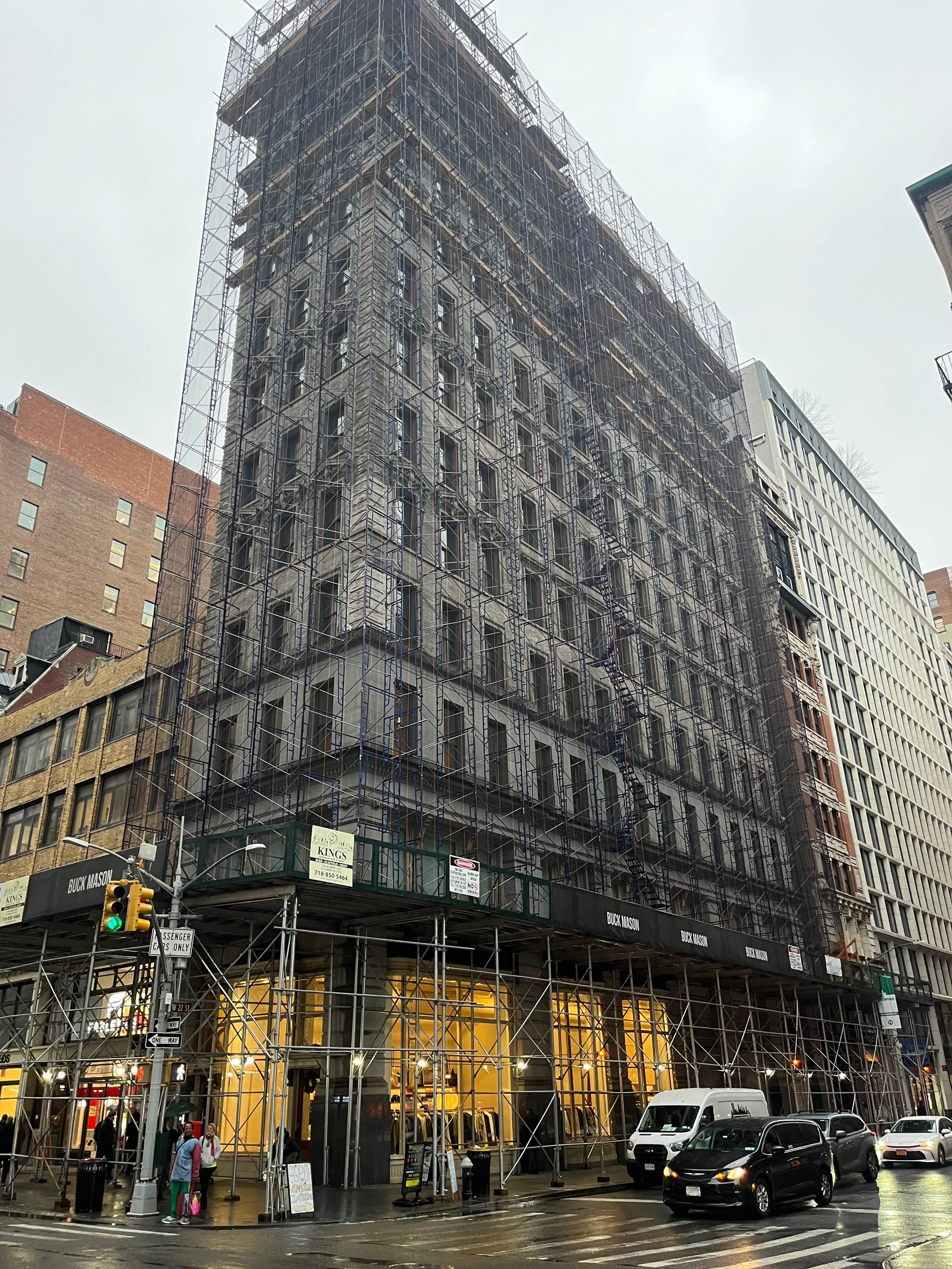 A tall building under construction surrounded by scaffolding, with people and cars on the street in front.