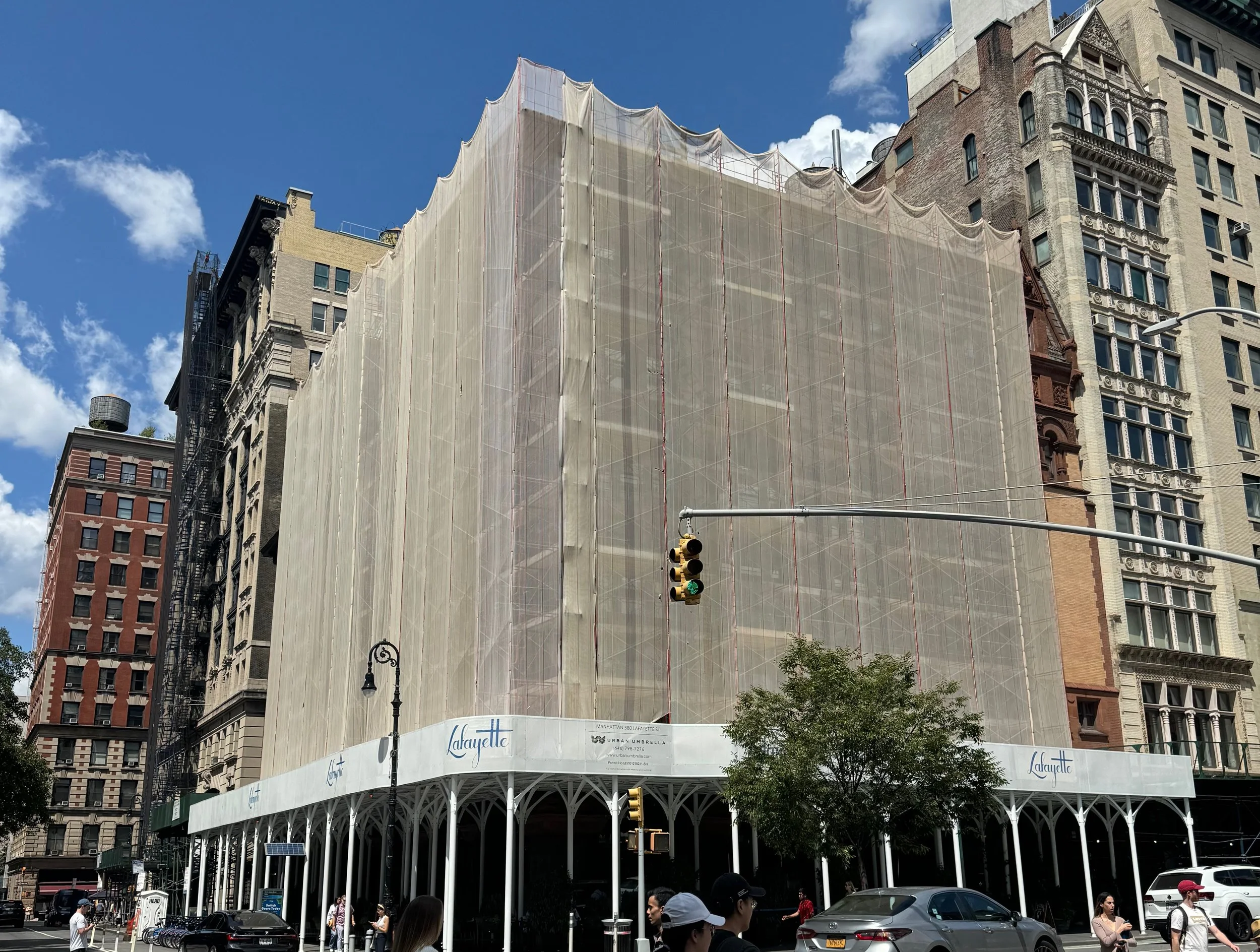 City street scene with a building under renovation, covered by beige scaffolding and wrapped in protective netting, with pedestrians and cars in the foreground.