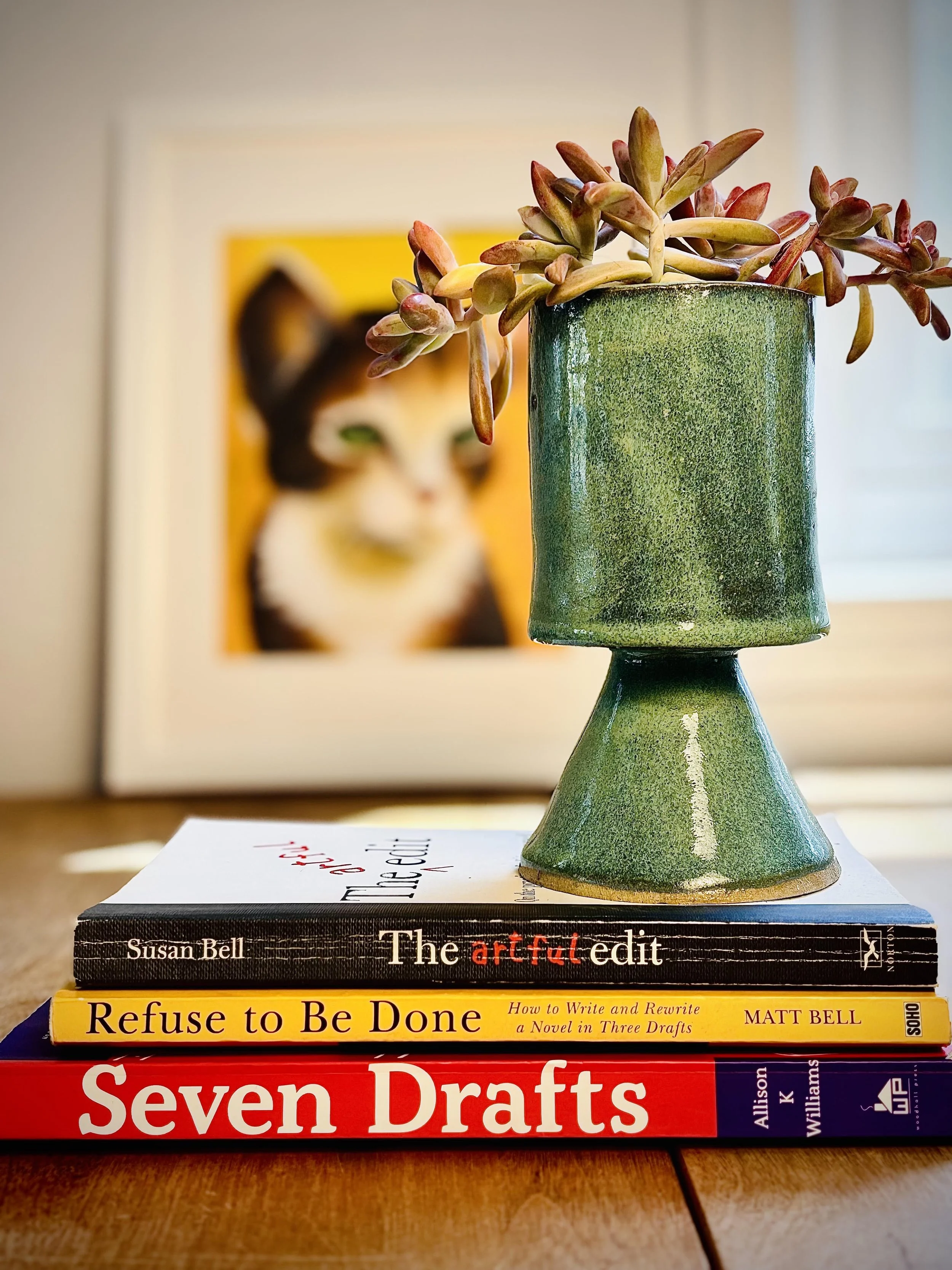 A small succulent plant in a green ceramic pot is placed on a stack of books. The books include "The Artful Edit" by Susan Bell, "Refuse to Be Done" by Matt Bell, and "Seven Drafts" by Allison Williams. In the background, there is a blurred image of a cat portrait with a yellow background.
