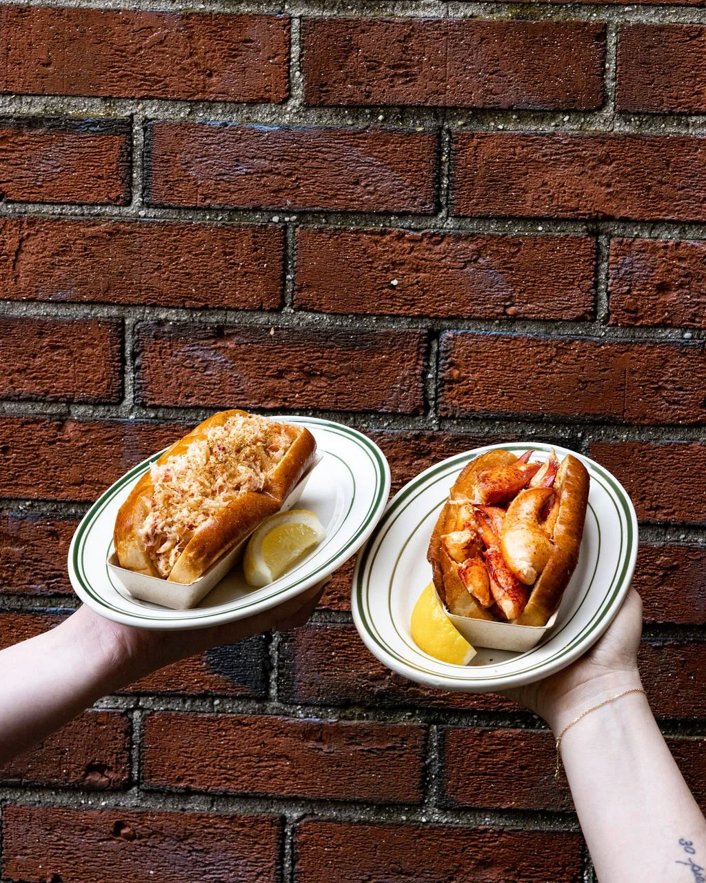 One hand holding a plated lobster roll, the other holding a plated crab roll