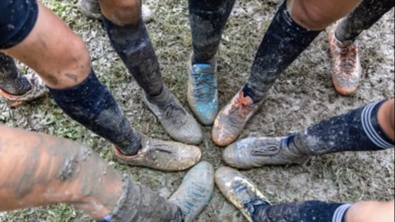 Group of muddy soccer or rugby players standing in a circle, showing their dirty shoes and socks on a grassy field.