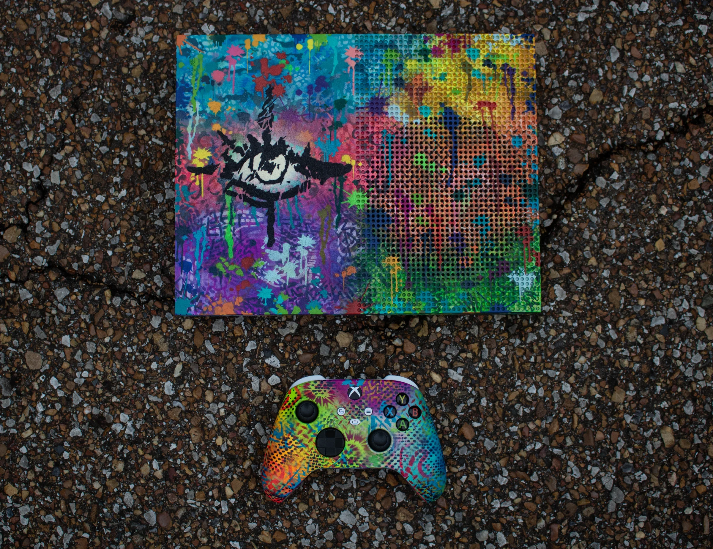 Colorful graffiti-themed Xbox controller and matching console on gravel ground, with the controller in the foreground and the console behind it.