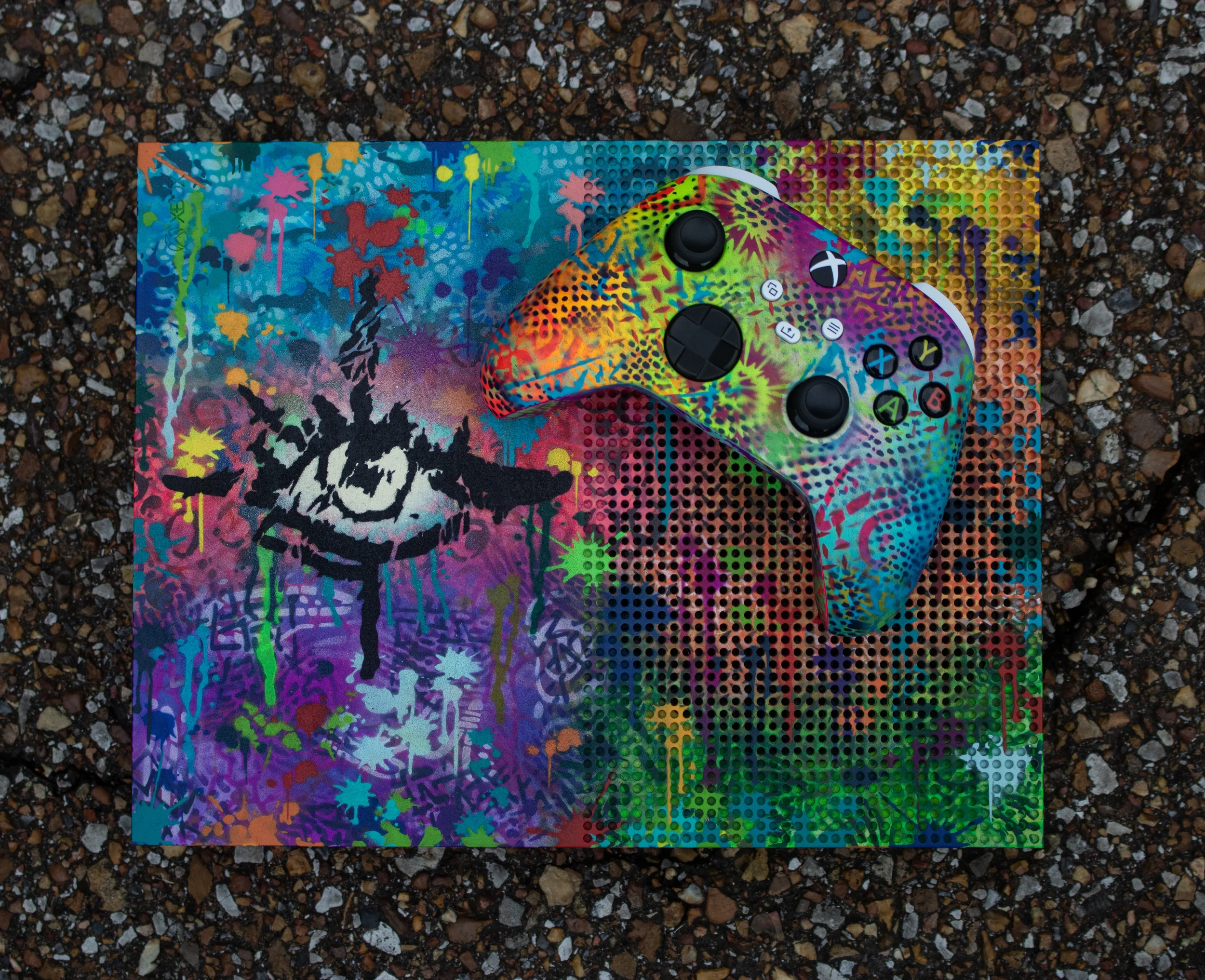 A colorful, graffiti-style painted Xbox controller resting on a matching graffiti-painted box, placed on a gravel surface.
