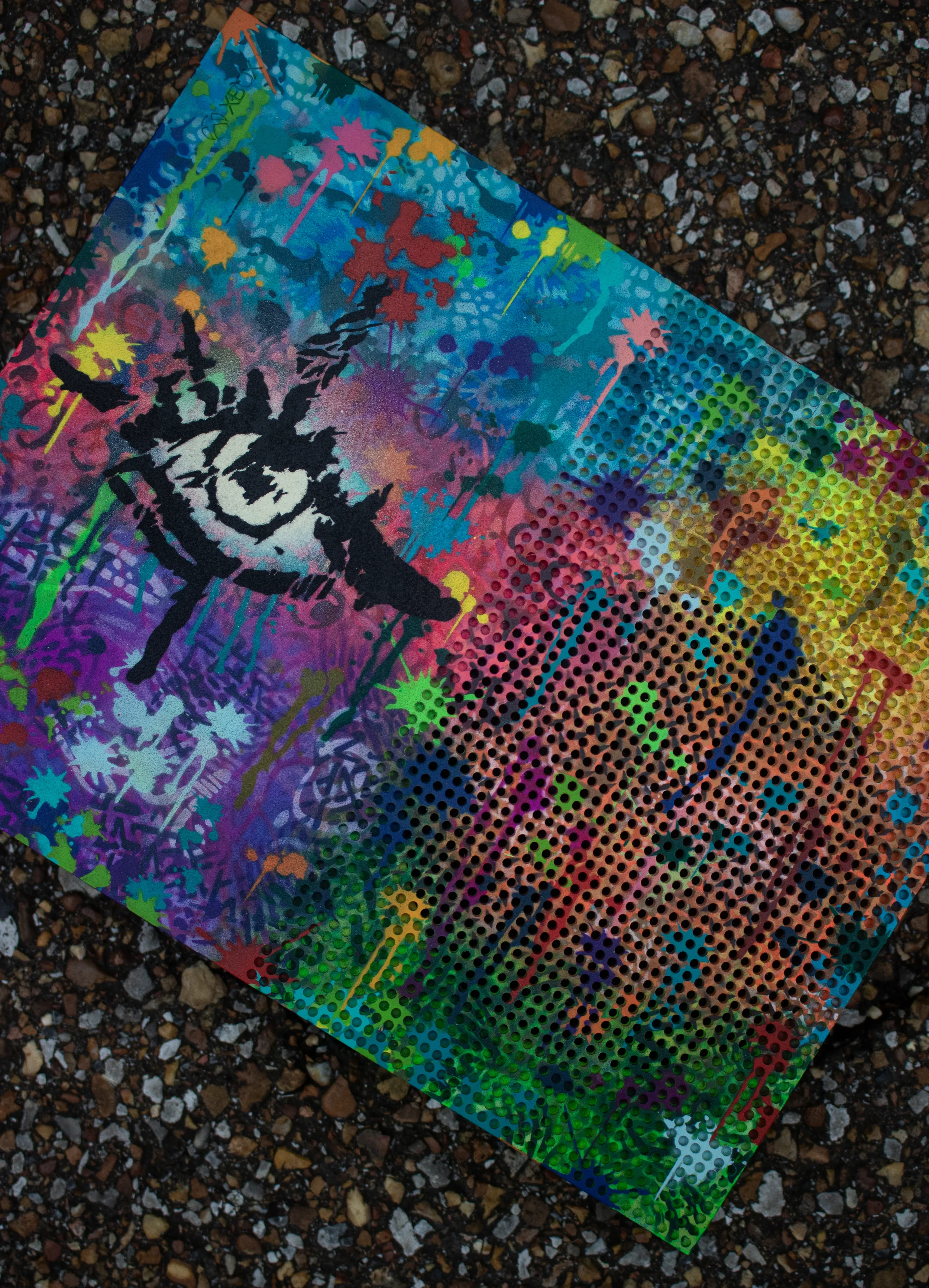 Colorful graffiti-style artwork on a perforated sheet resting on a gravel surface.