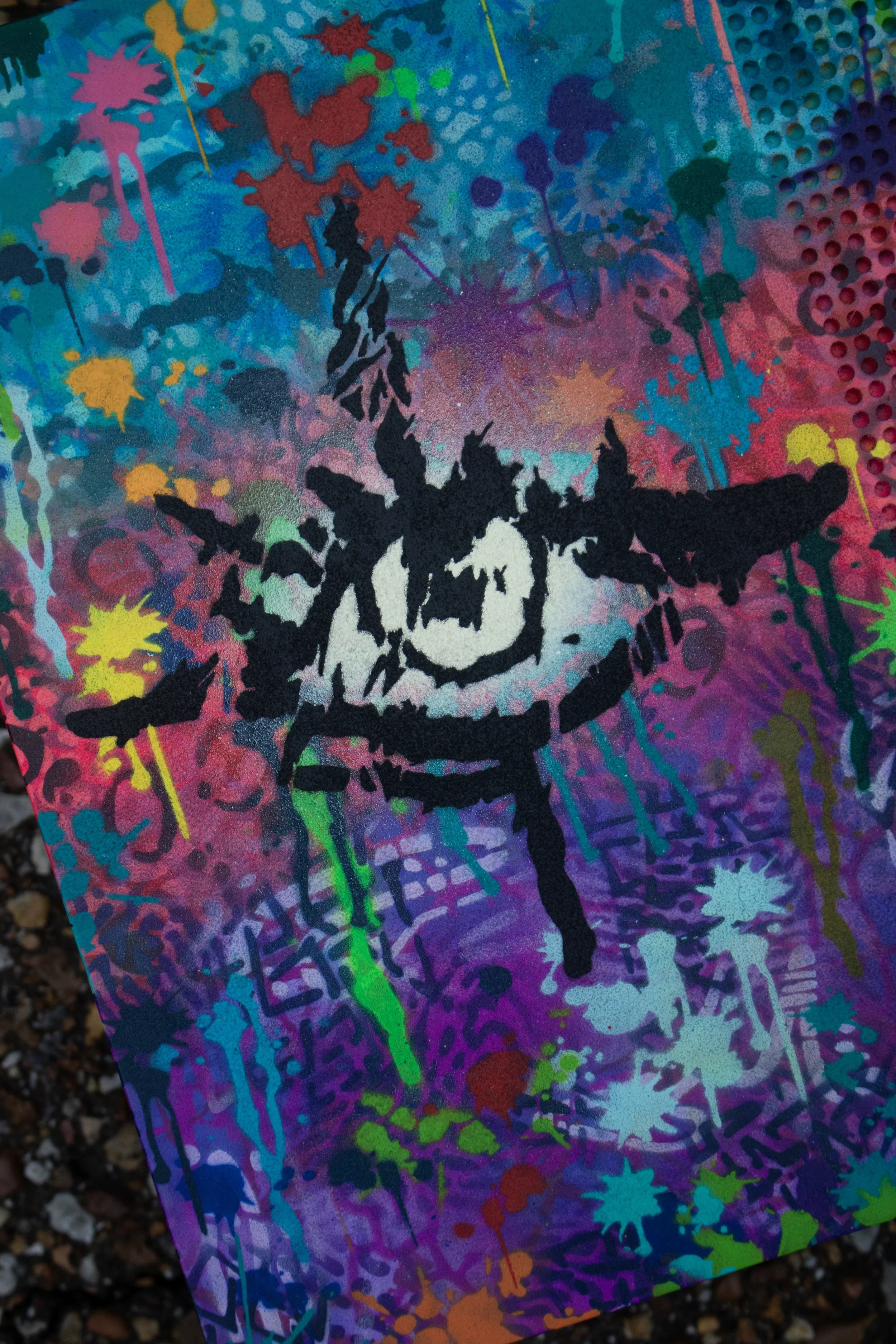 A colorful abstract graffiti art with splashes of pink, yellow, green, blue, purple, and orange paint, featuring a black stencil of a child's face with a stern expression.