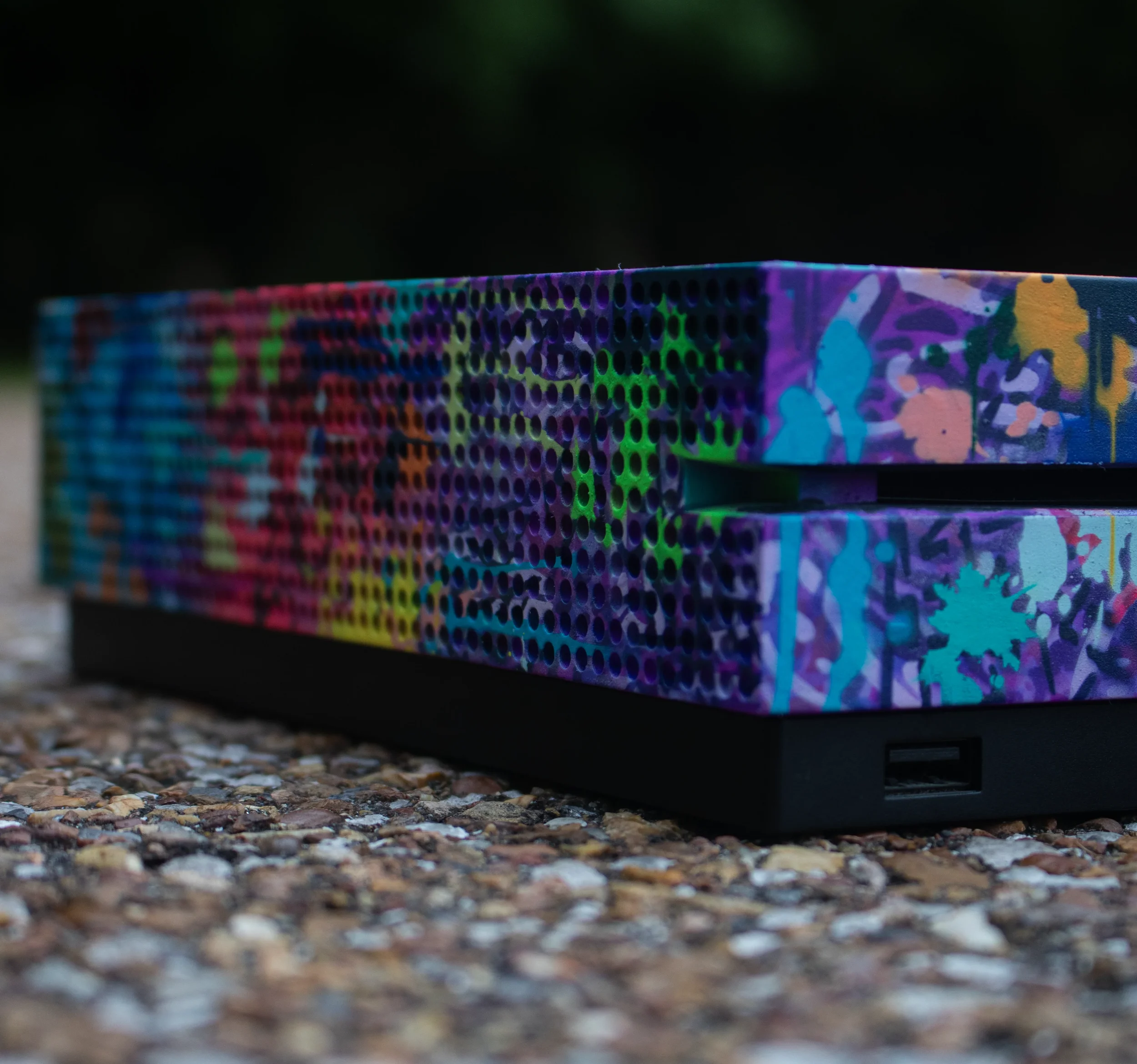 Close-up of a colorful skateboard deck with abstract graffiti art, resting on a gravel surface with a dark background.