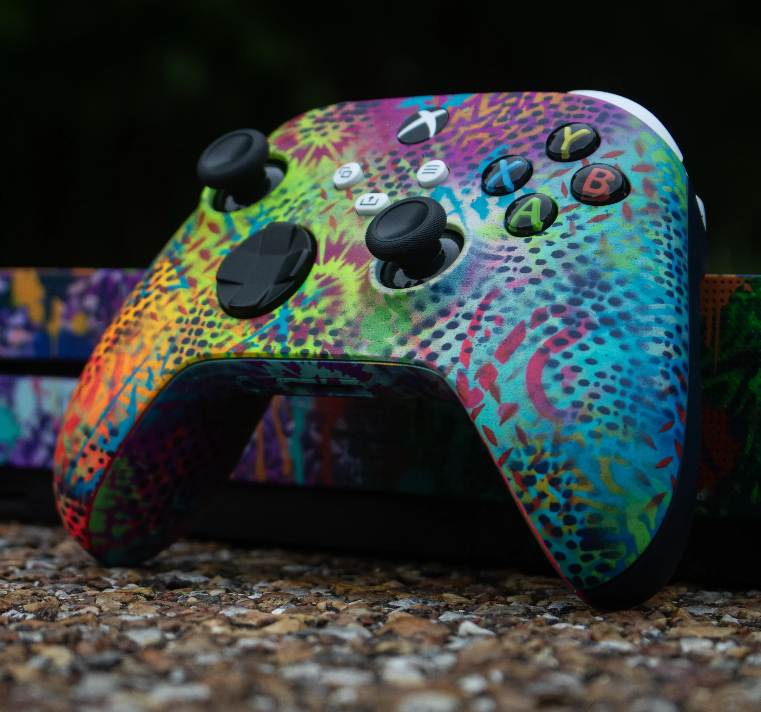 Close-up of a colorful, paint-splattered guitar body with black knobs and white buttons, resting on a gravel ground with a blurred dark background.