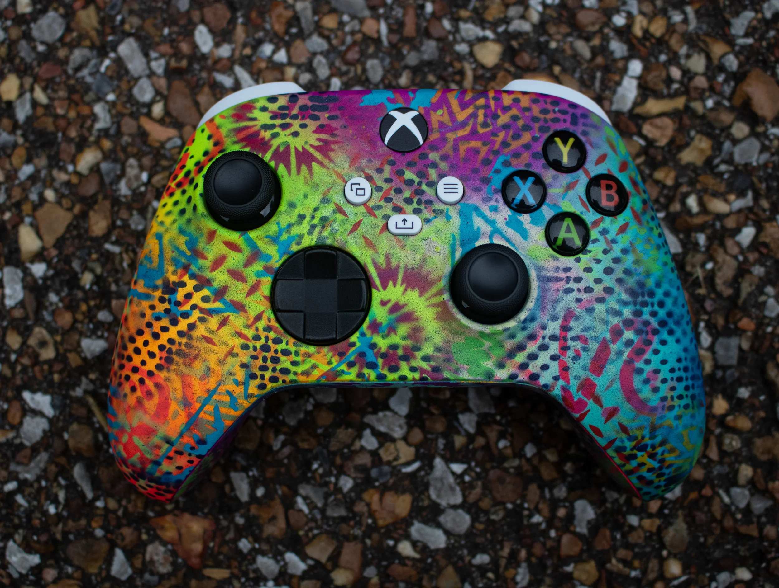 A colorful Xbox controller with a splatter paint design, lying on a gravel surface.