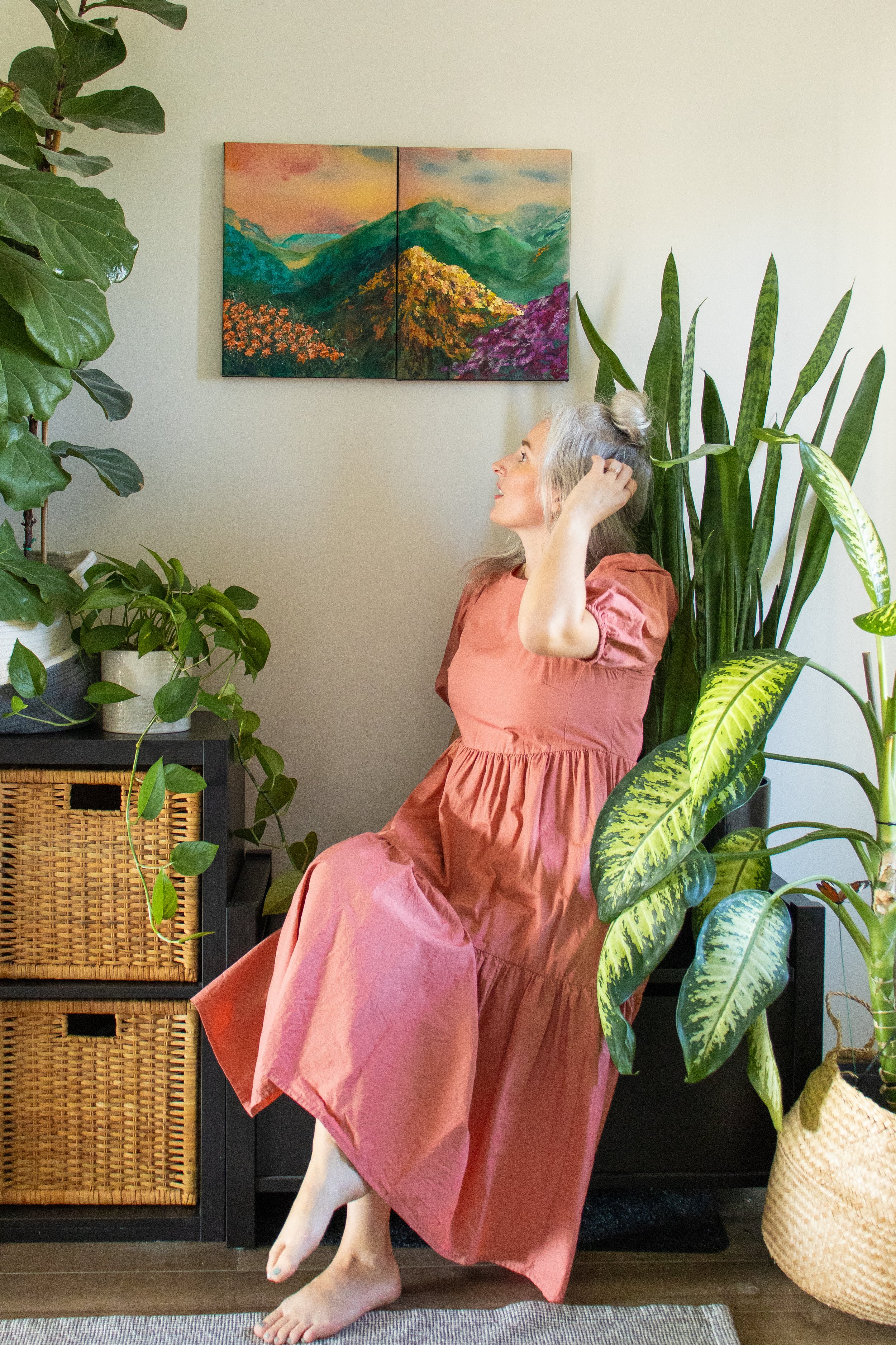 Artist, Rebekah Victoria sitting amongst green foliage and indoor plants in front of her paintings of a beautiful landscape that is dreamy and floral at sunset.