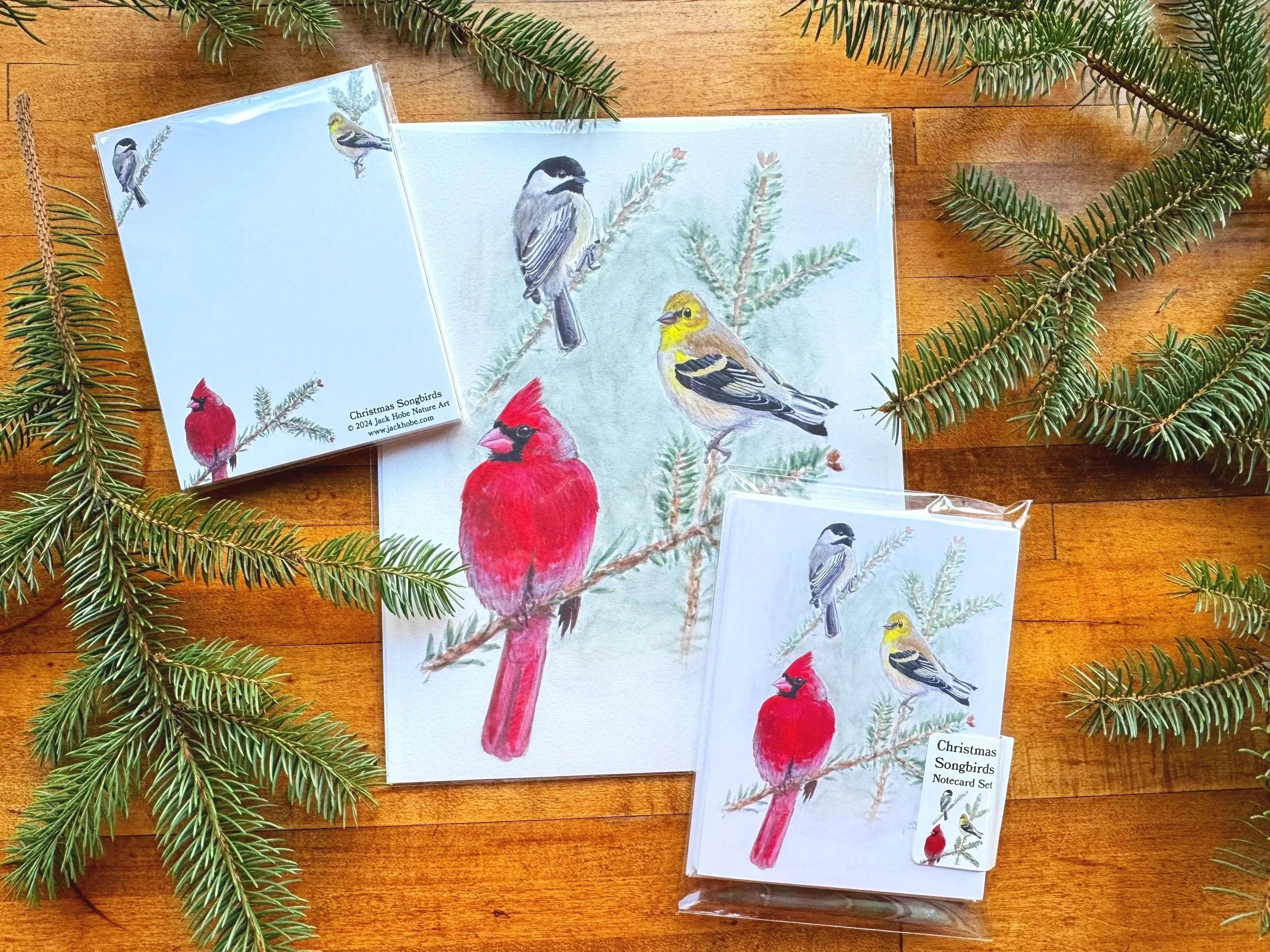 Set of Christmas notecards and sticker with watercolor illustrations of various songbirds, including a cardinal, chickadee, and goldfinch, on a wooden surface decorated with pine branches.