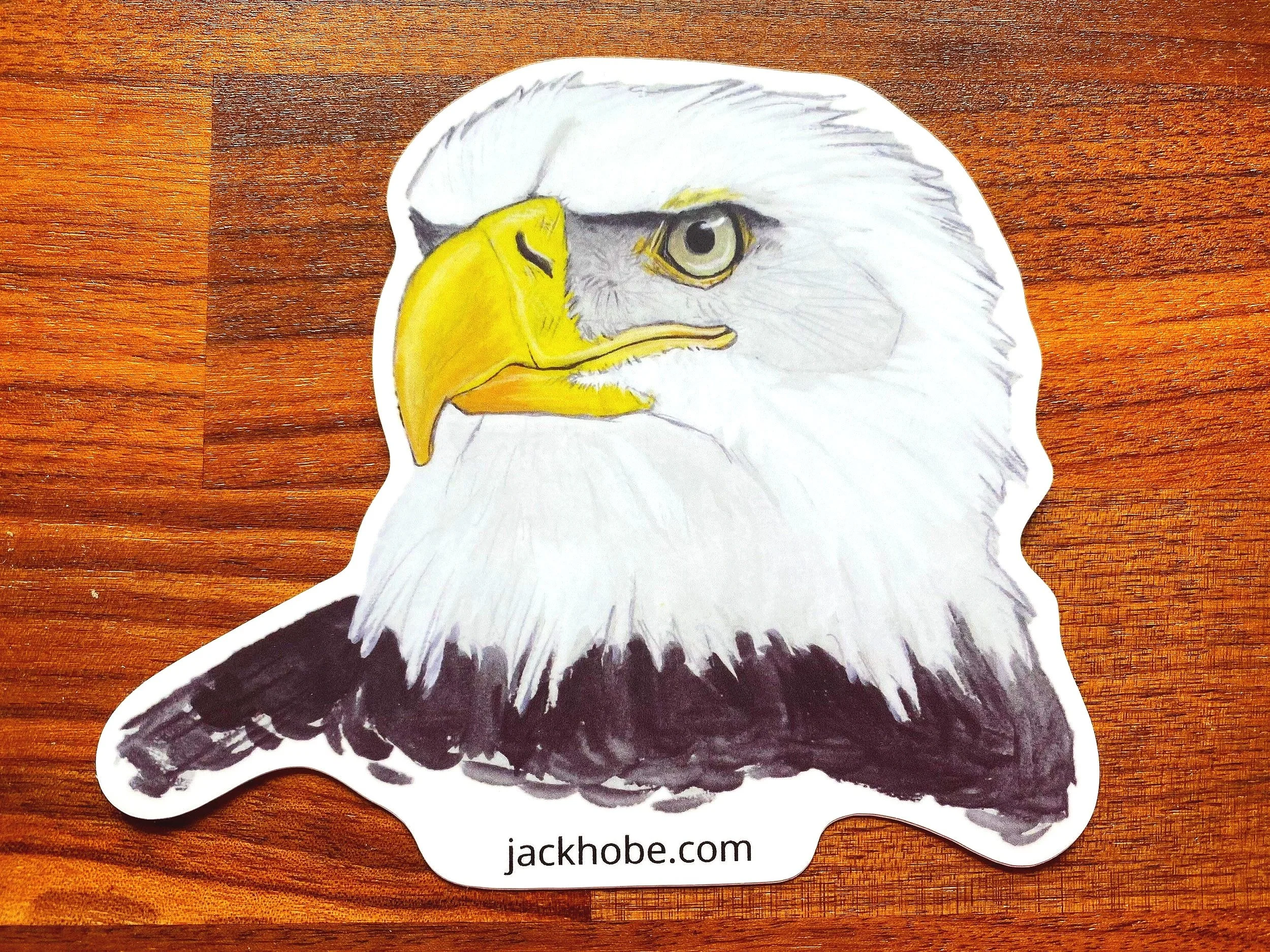 Illustration of a bald eagle's head with a yellow beak and white feathers, on a wooden surface, with the text "jackhobe.com" at the bottom.