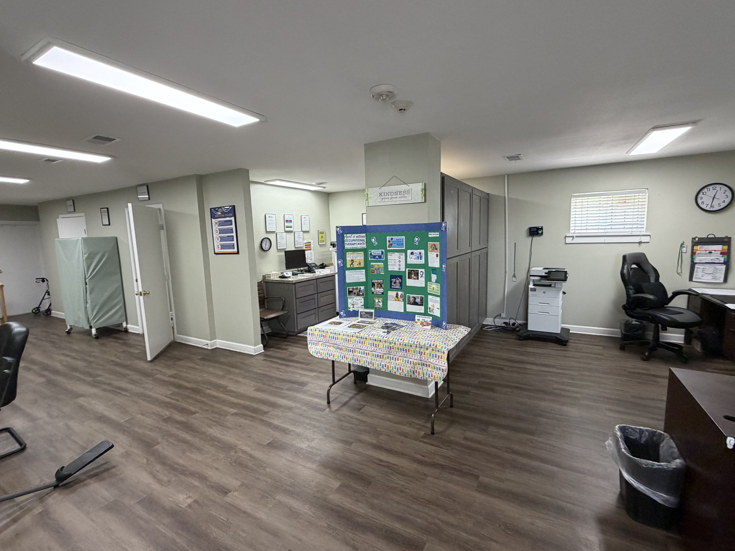 An empty office or clinic room with a bulletin board, tables, chairs, computer, copier, clock, and a window with blinds.
