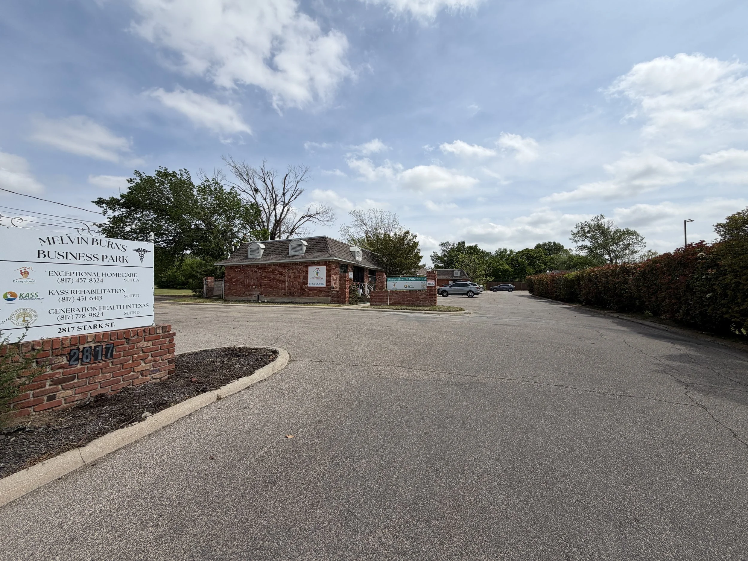 View of Melvin Burns Business Park with a brick building, parking lot, and signs listing various businesses including Exceptional Homecare, KASS Revalidation, Generation Health TN, under a partly cloudy sky with trees in the background.