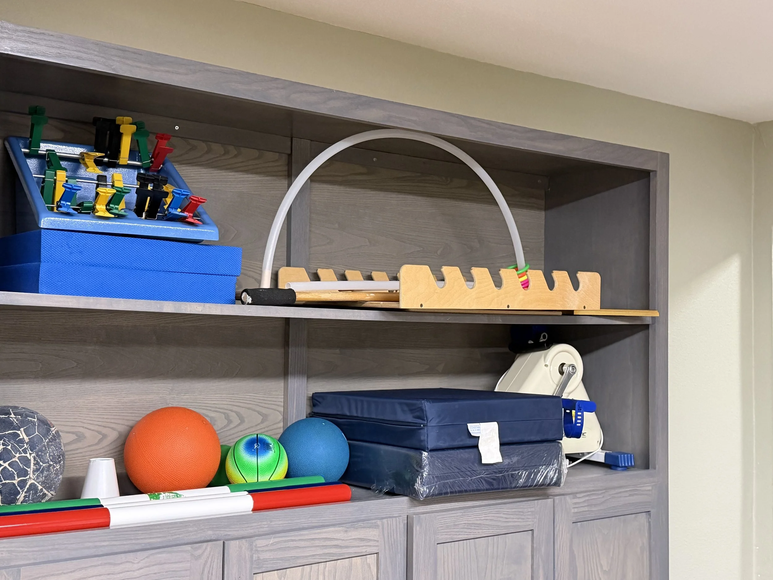 Shelves with sports and therapy equipment including foam balls, a balance board, a foam roller, a small hockey stick, and various small parts for exercises.