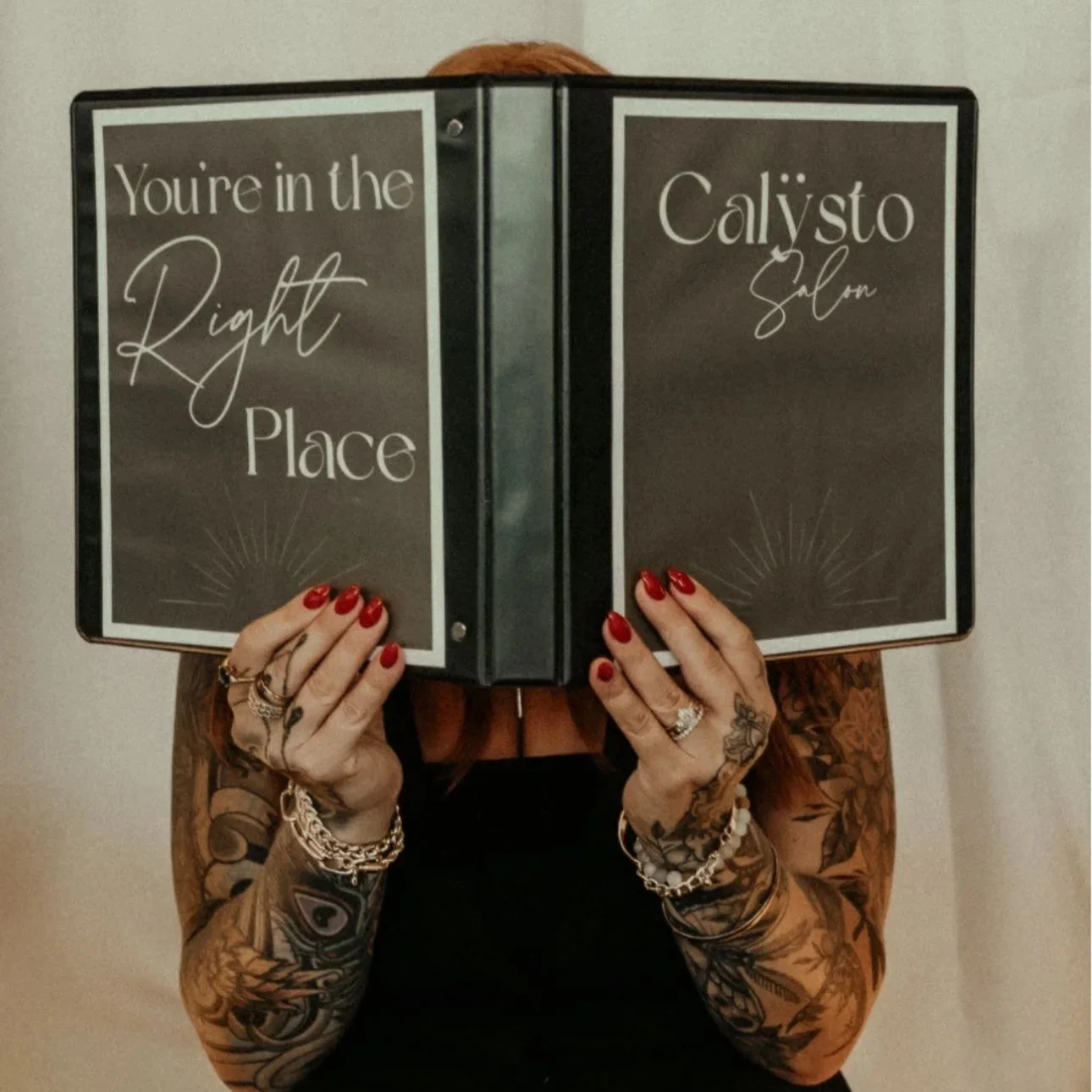 Person with red hair and tattoos holding a black menu or sign with the text 'You're in the Right Place' and 'Calysto Salon' written on it.