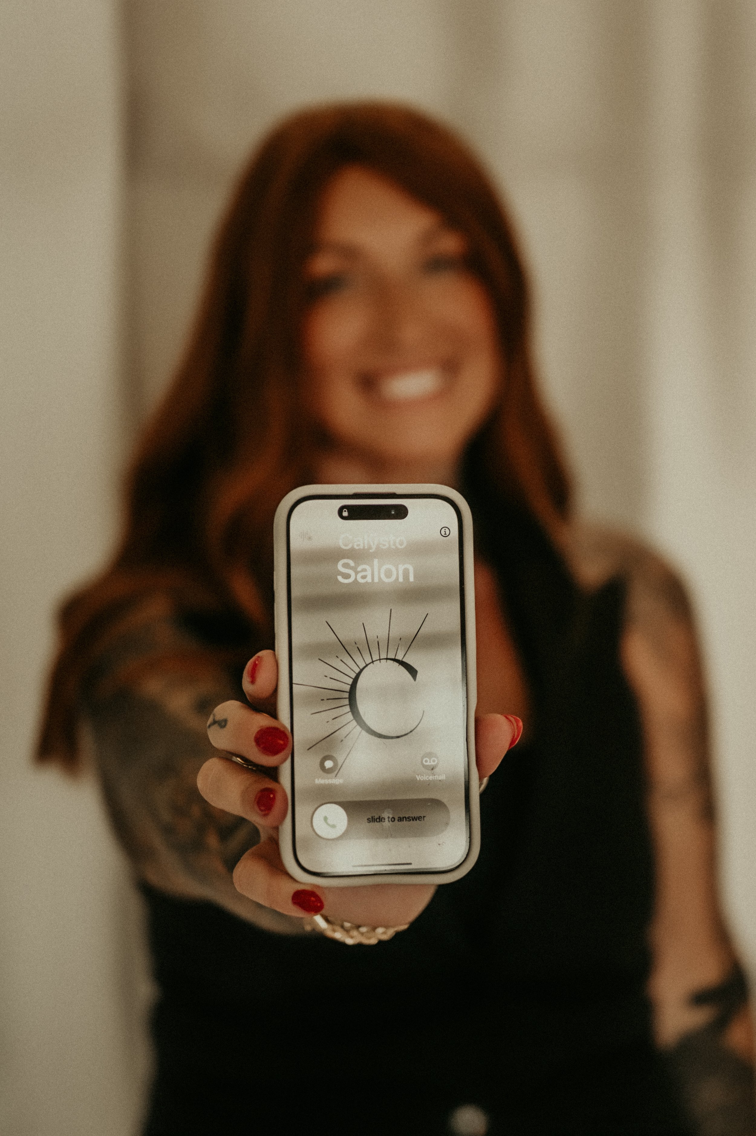Owner Jamie Harrington holding a smartphone showing a call from 'Calysto Salon' Calysto Salon the best place for hair color : blonde, balayage, dimensional lived in color, hair extensions, wigs, mesh integration topper
