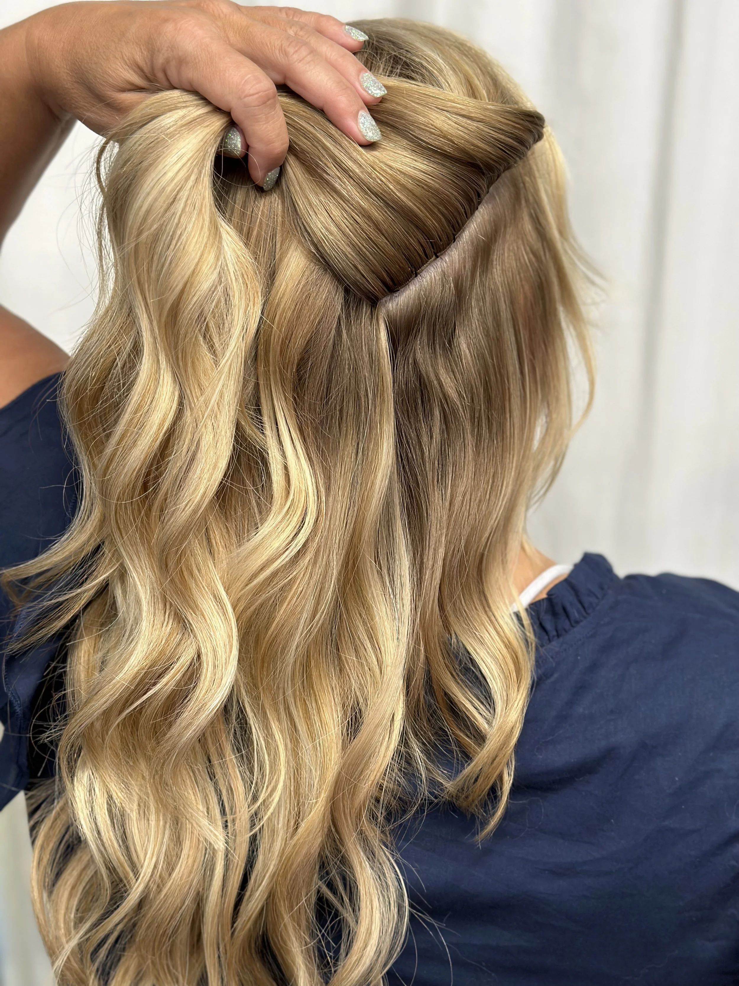 A hairstylist is arranging long, blonde, wavy hair, part of it being held up to showcase a section of the hair.